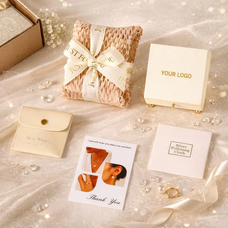 Elegant jewelry packaging set on shimmery fabric, including a woven cushion wrapped with a ribbon, a cream jewelry box labeled 'YOUR LOGO', a small pouch with 'Best Wishes', a silver polishing cloth packet, a thank you card, and a gold clasp accessory.
