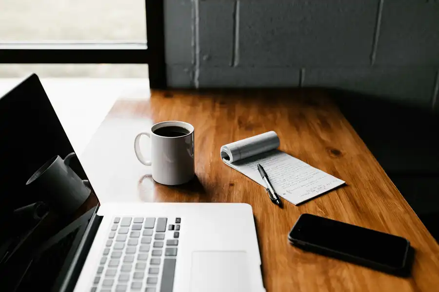 Workspace with a laptop, white coffee mug, notepad with handwritten notes, pen, and a smartphone on a wooden table near a window.