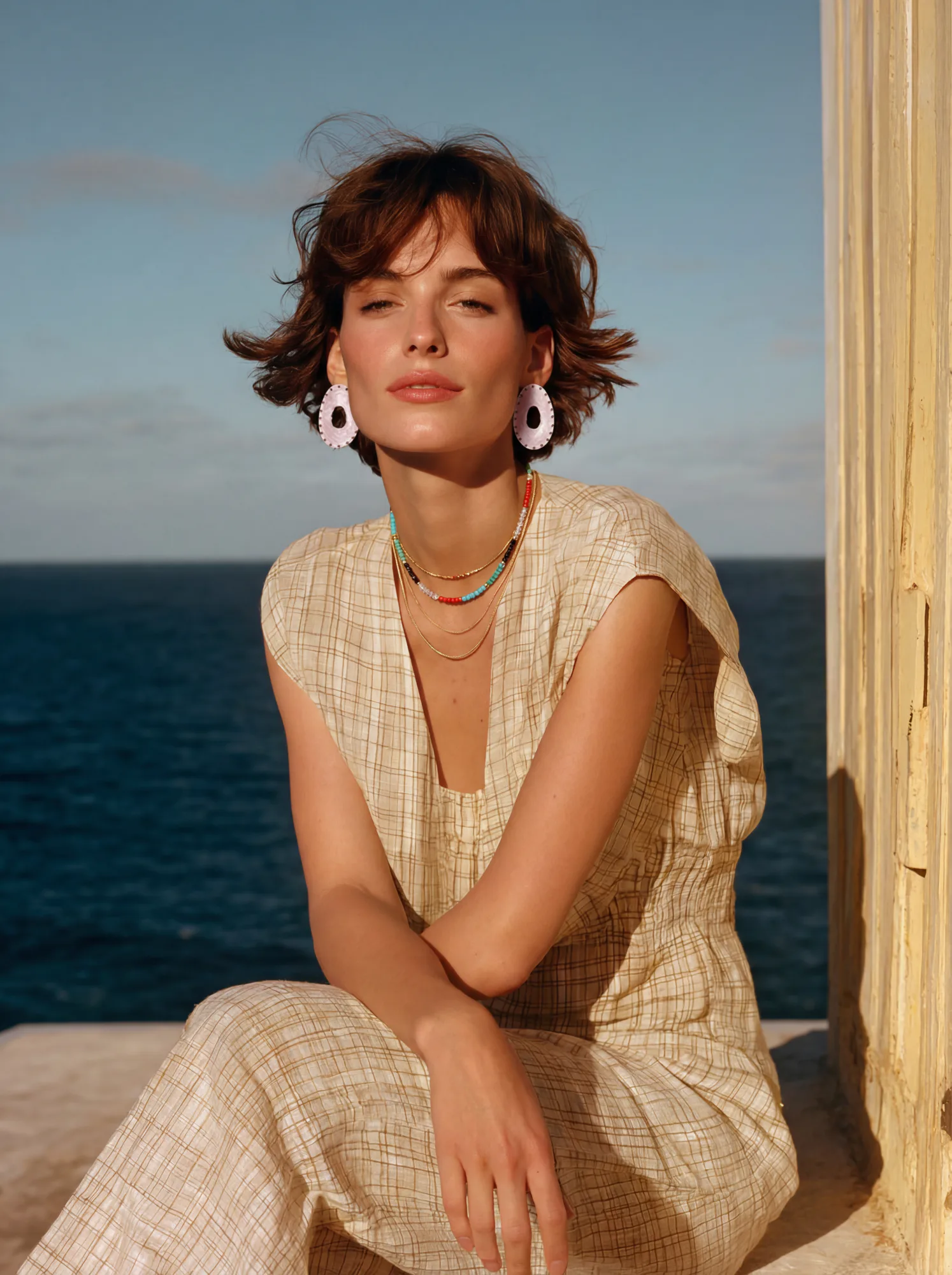 Woman with short brown hair wearing a beige plaid dress and colorful necklaces seated by the ocean.
