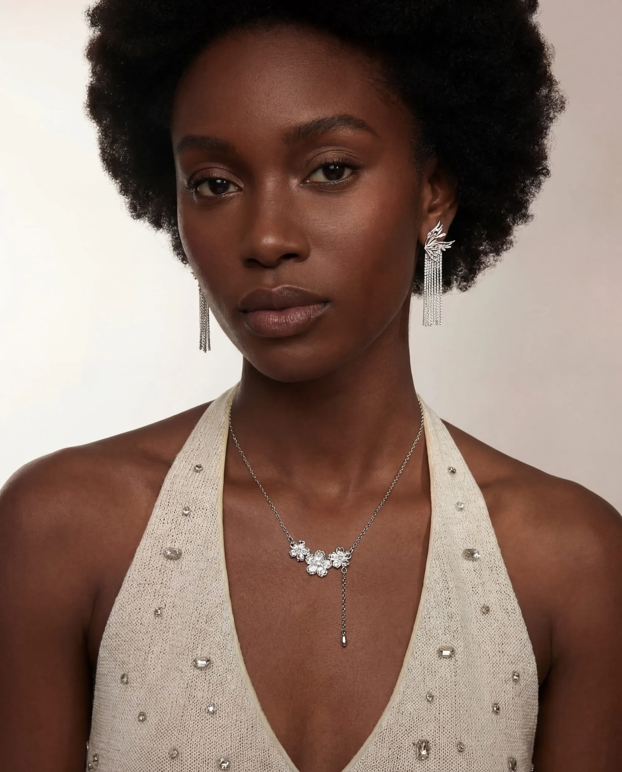 Woman with natural afro hairstyle wearing silver dangling earrings and a silver necklace with floral-shaped gems, dressed in a beige halter top embellished with small stones.