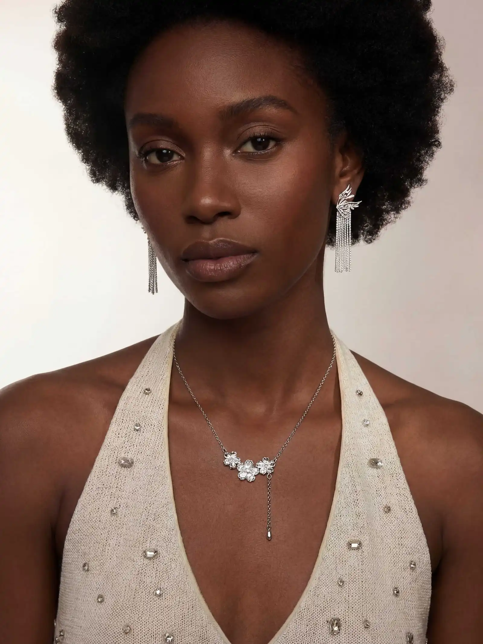 Woman with natural afro hairstyle wearing silver dangling earrings and a silver necklace with floral-shaped gems, dressed in a beige halter top embellished with small stones.