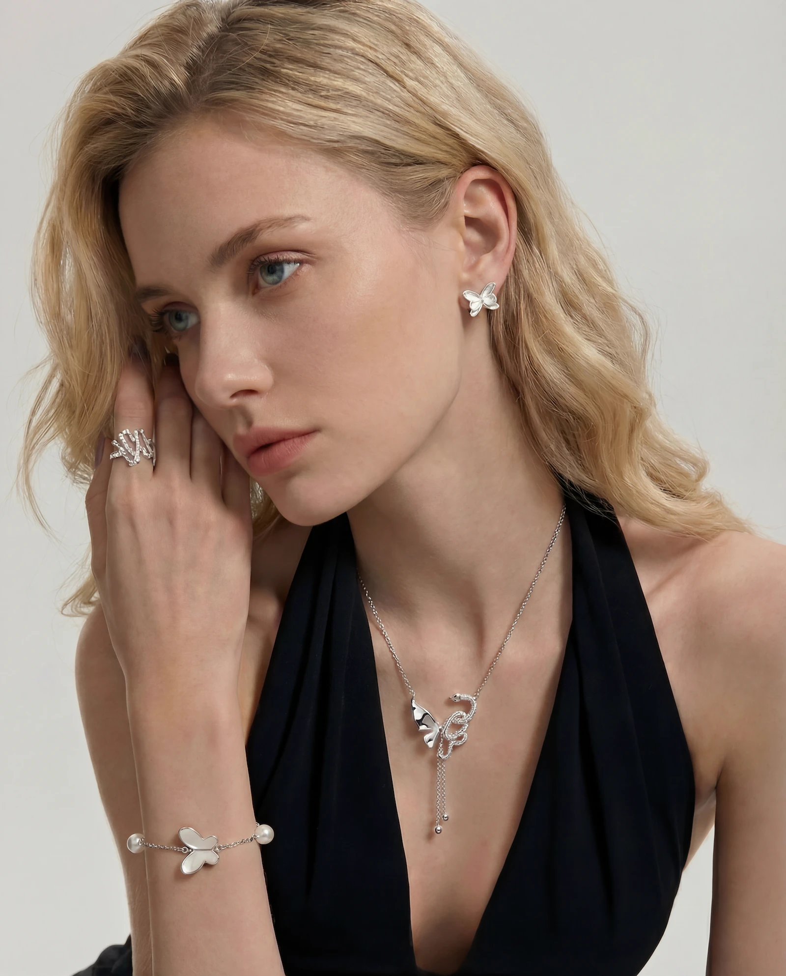 Blonde woman wearing silver butterfly-shaped earrings, a matching butterfly bracelet with pearls, a silver ring, and a necklace with butterfly and snake pendants.