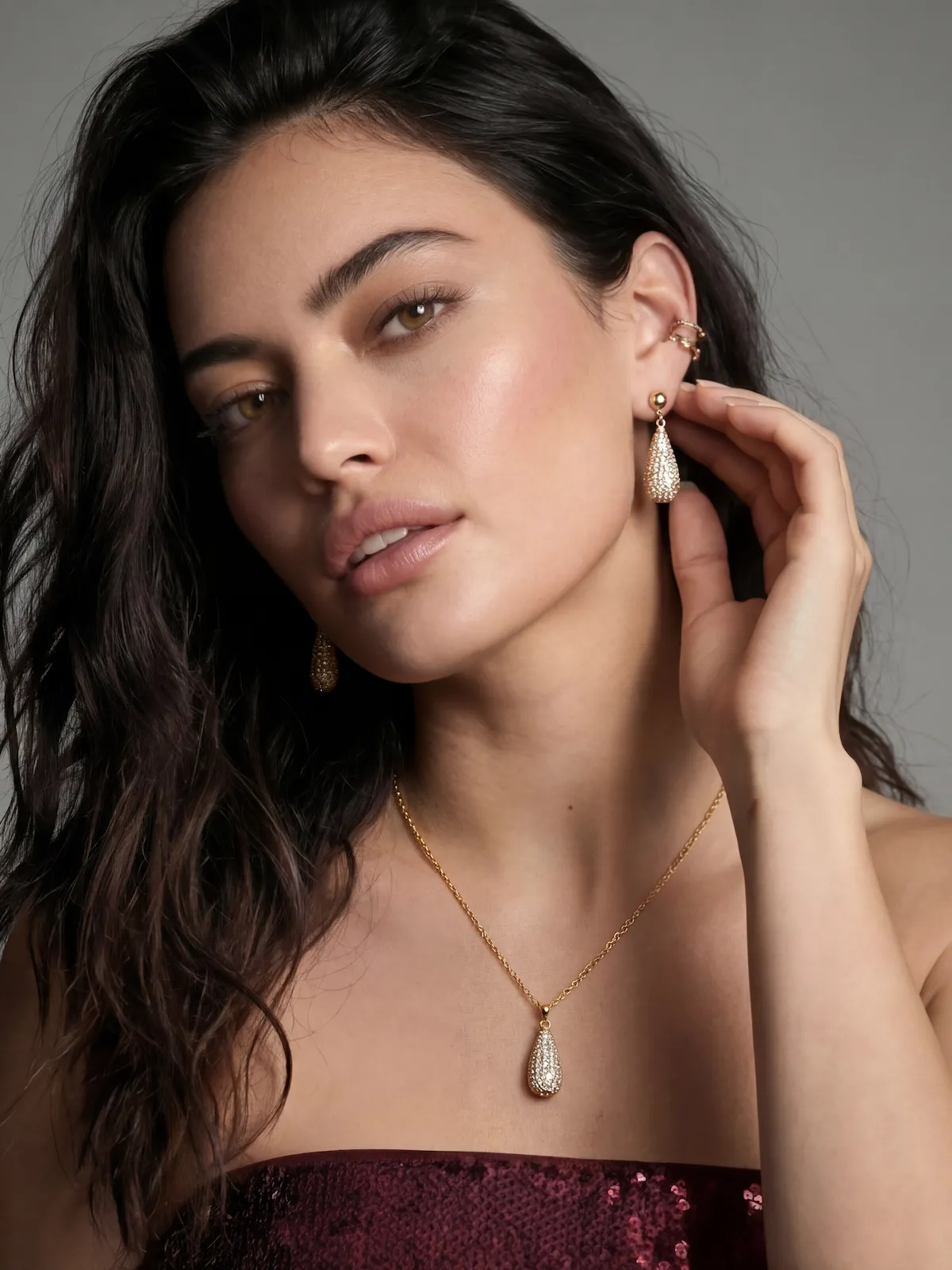 Woman with long dark hair wearing gold teardrop earrings and matching necklace against a gray background.