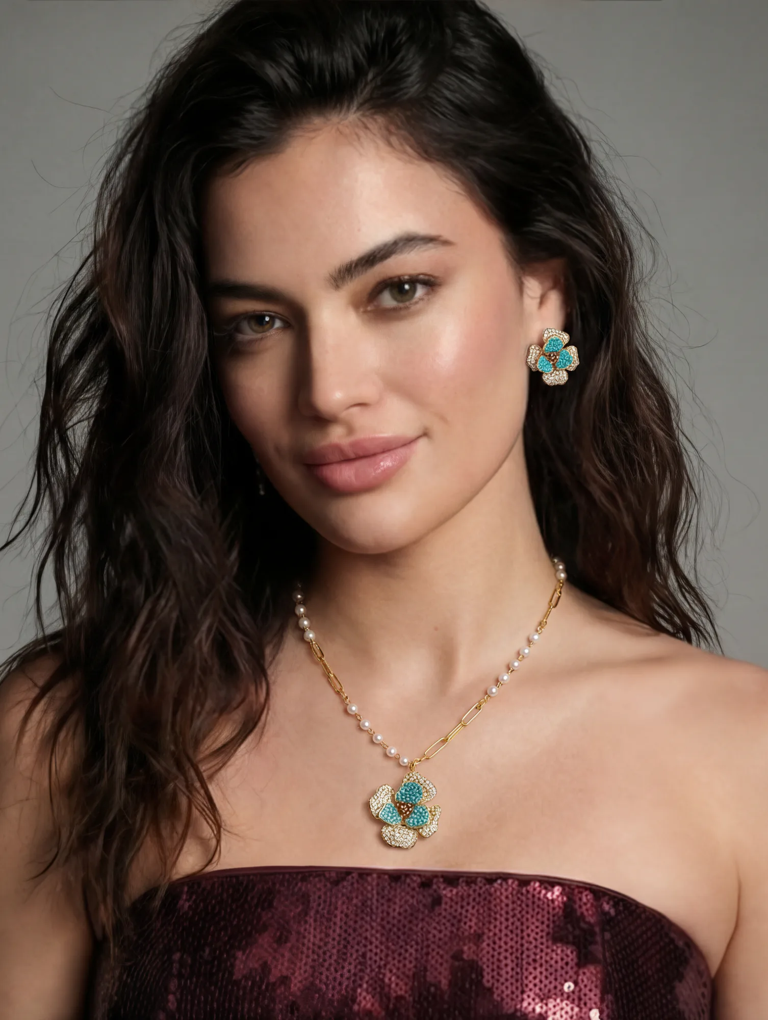 Woman with long dark hair wearing gold teardrop earrings and matching necklace against a gray background.