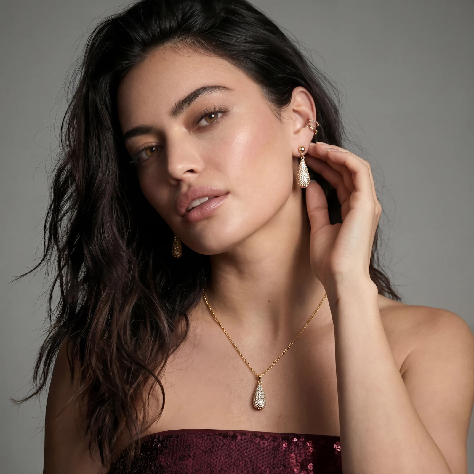 Woman with long dark hair wearing gold teardrop earrings and matching necklace against a gray background.