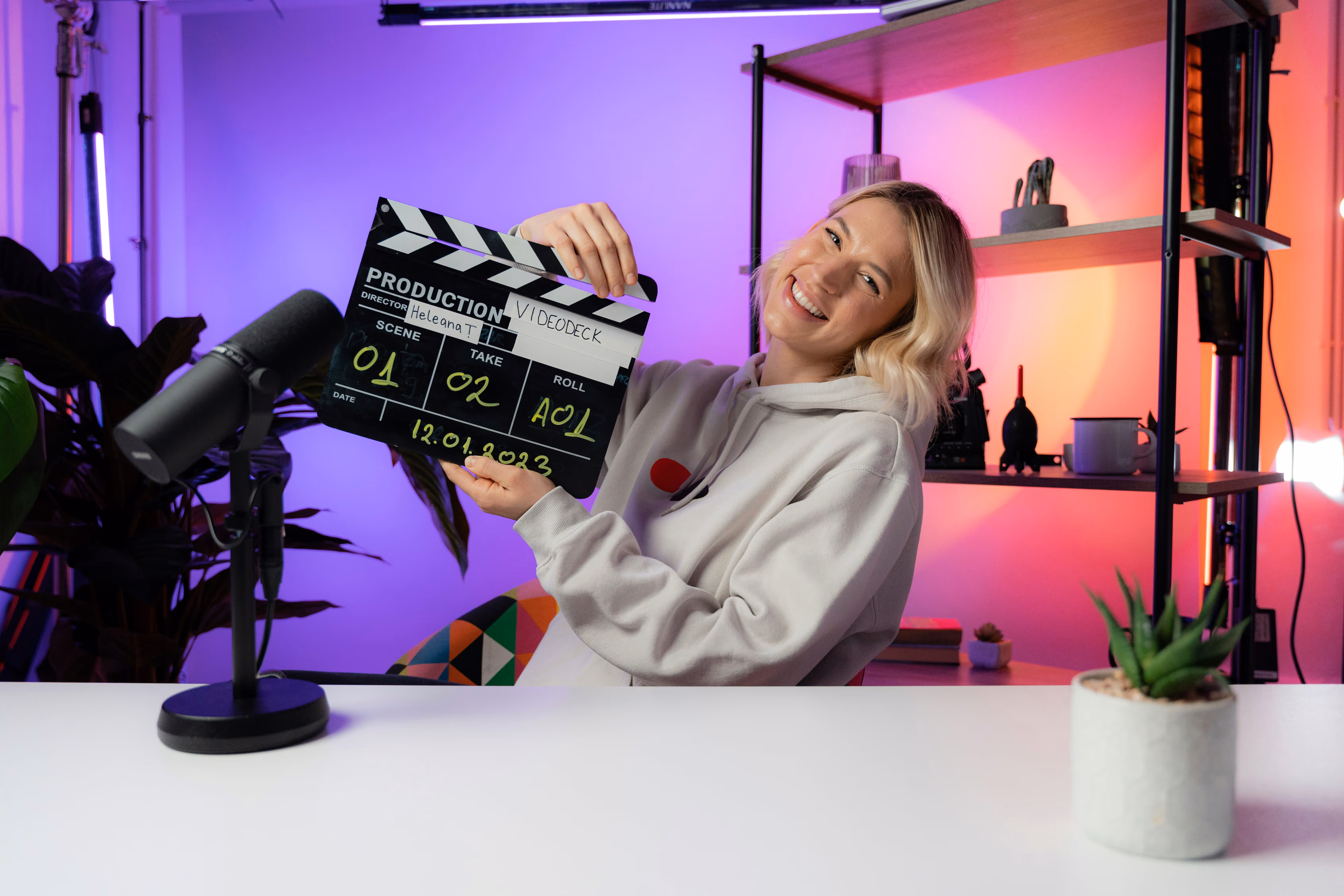 Smiling woman in a hoodie holding a film clapperboard in a colorful studio with a microphone and shelves in the background.