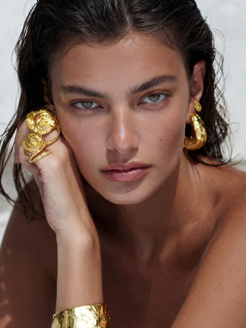Close-up of a woman with wet hair wearing large textured gold earrings, rings, and a cuff bracelet.