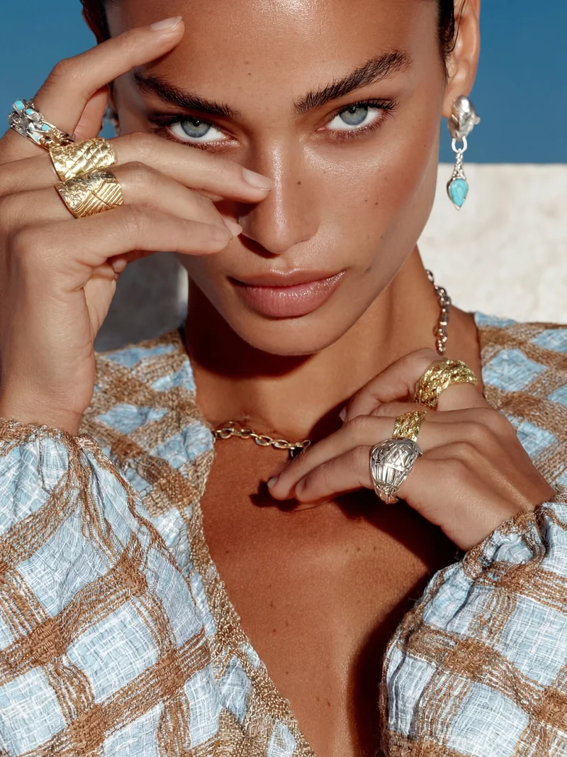 Close-up of a woman with blue eyes wearing gold and silver rings, a blue patterned top, and turquoise earrings.