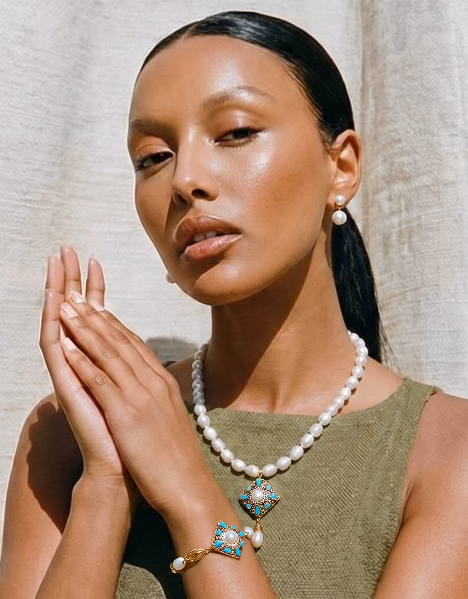 Close-up of a woman with smooth skin and dark hair in a ponytail wearing pearl earrings, a pearl necklace with a decorative turquoise and gold pendant, and a matching bracelet, posing with hands gently clasped against a neutral fabric backdrop.