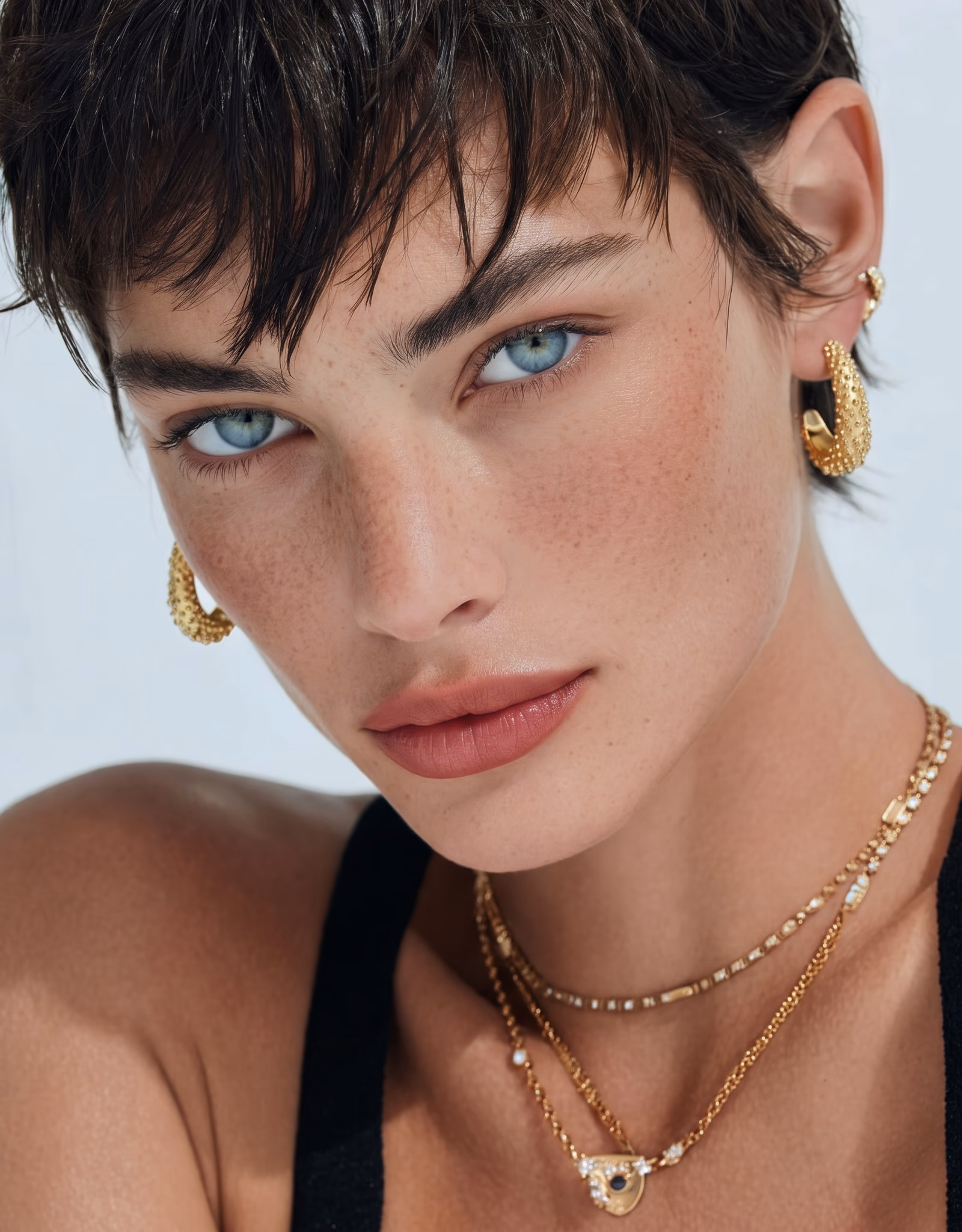 Close-up portrait of a person with short dark hair, striking blue eyes, and freckles, wearing gold hoop earrings and layered gold necklaces, against a light background.