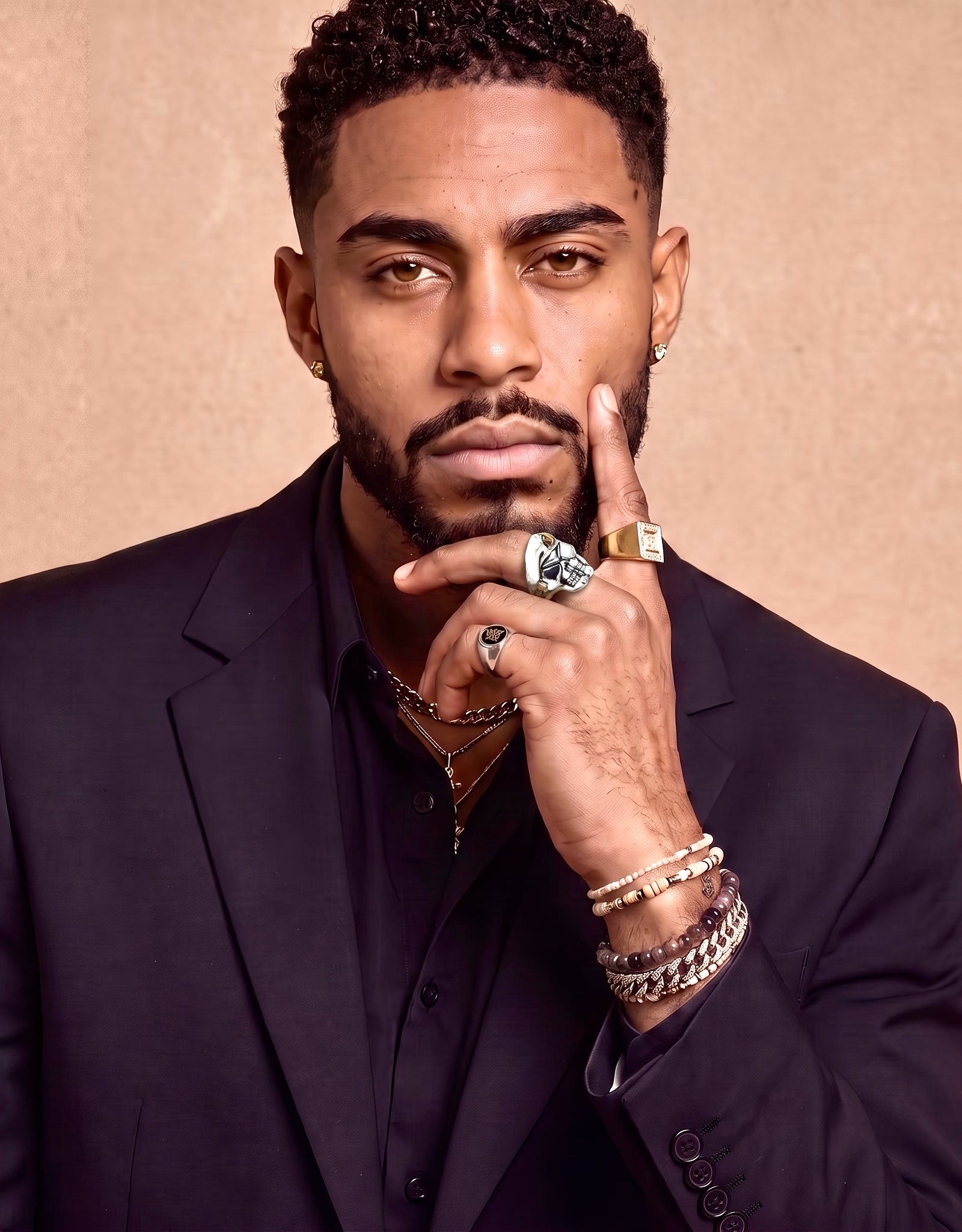 A man with short curly hair and a beard wearing a black suit jacket and shirt, accessorized with multiple rings, bracelets, gold earrings, and layered necklaces, resting his chin on his left hand while looking directly at the camera against a beige background.