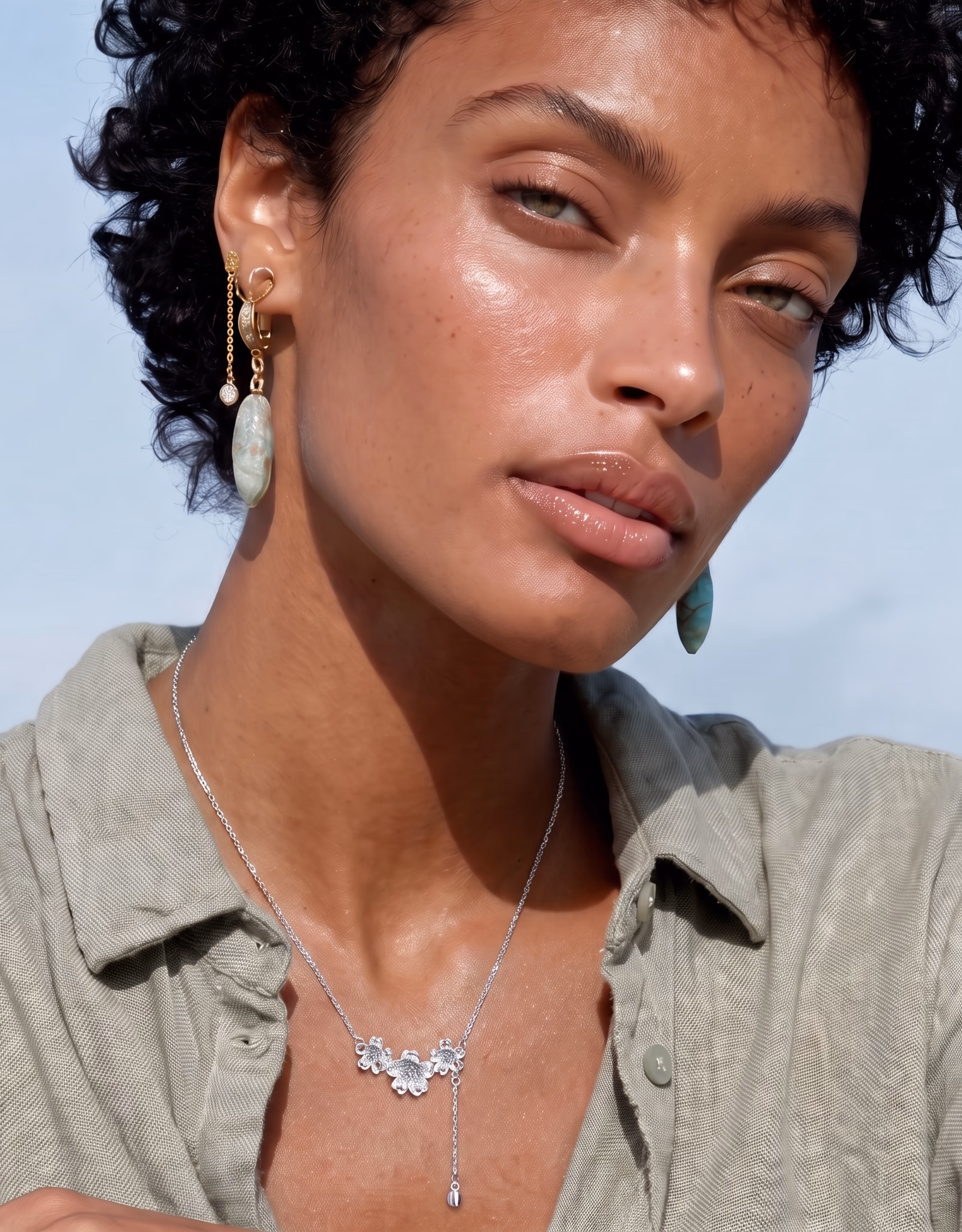 Close-up of a woman with short curly hair wearing dangling earrings with greenish stones and a silver floral necklace, dressed in an open-collared light green shirt against a light blue background.