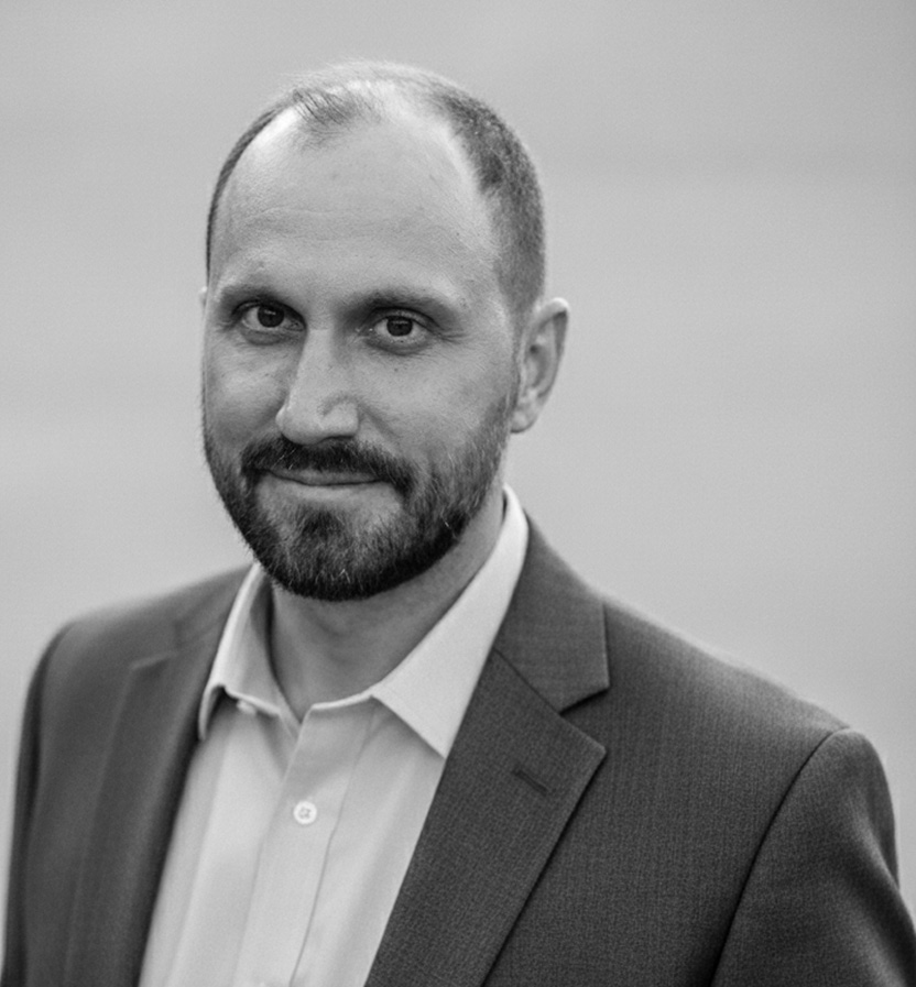 Black and white portrait of Jay Peterson with a beard wearing a suit jacket and collared shirt, smiling slightly.