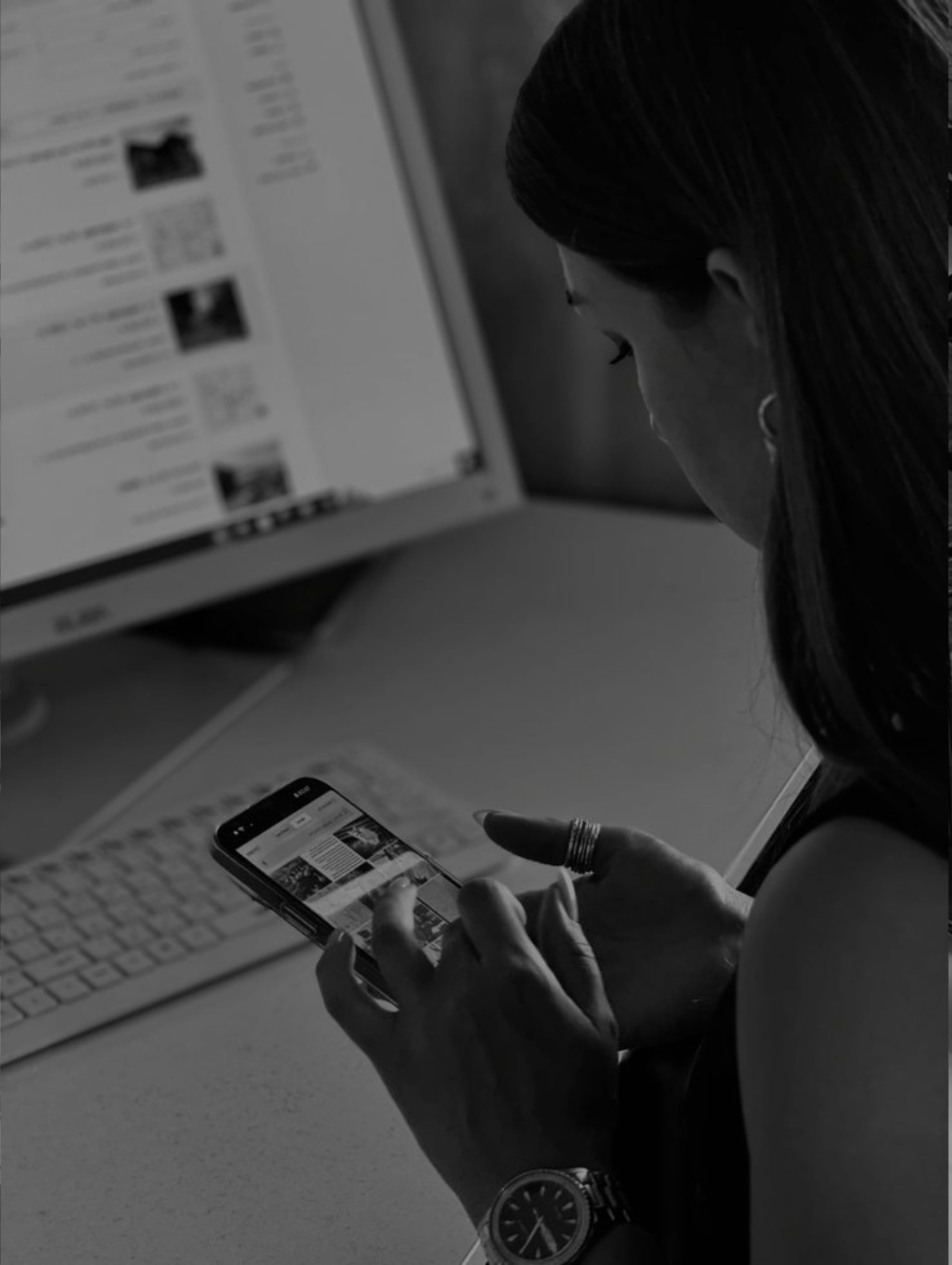 Woman using smartphone while sitting at desk with computer and keyboard in the background.