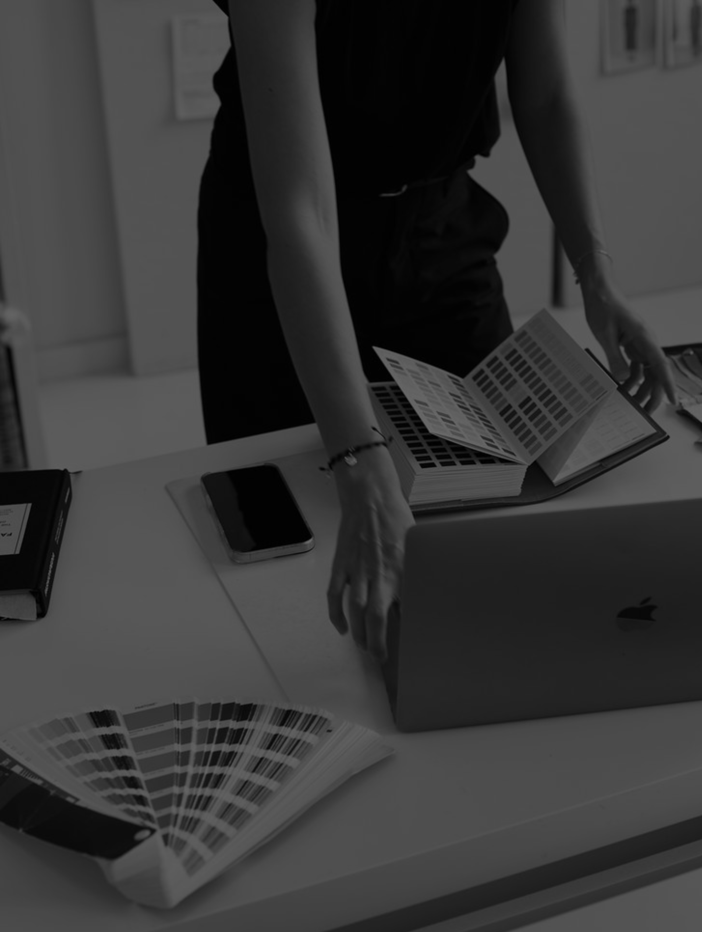 Person standing at a desk with color swatch books, a smartphone, and a laptop.
