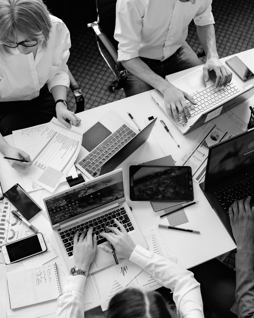 Overhead view of four people working on laptops and documents around a white office desk.