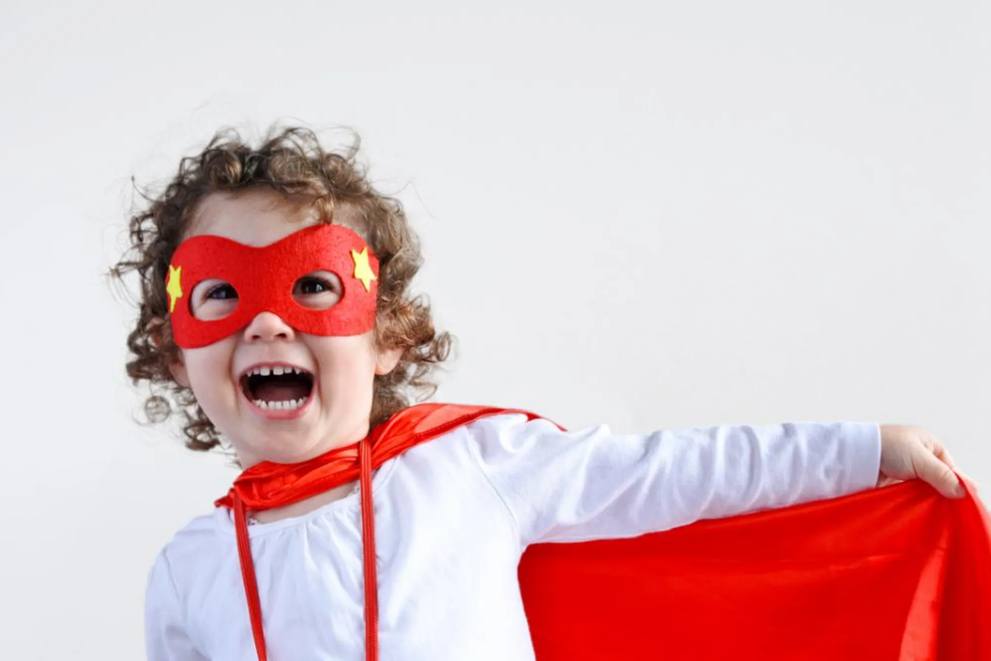 Smiling toddler wearing a red superhero mask and cape, holding the cape out to the side.