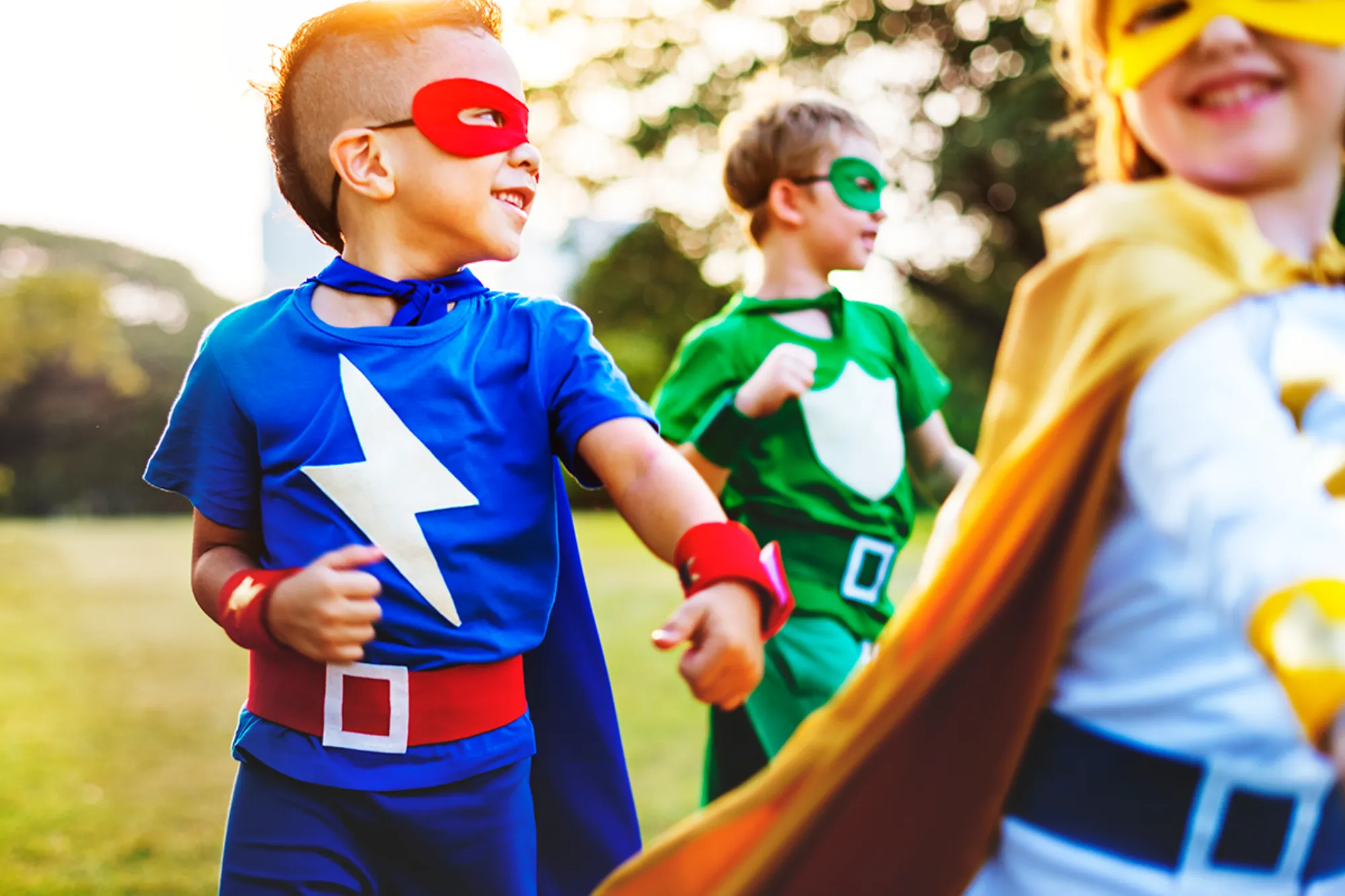 Three children playing outside dressed in colorful superhero costumes with masks and capes.