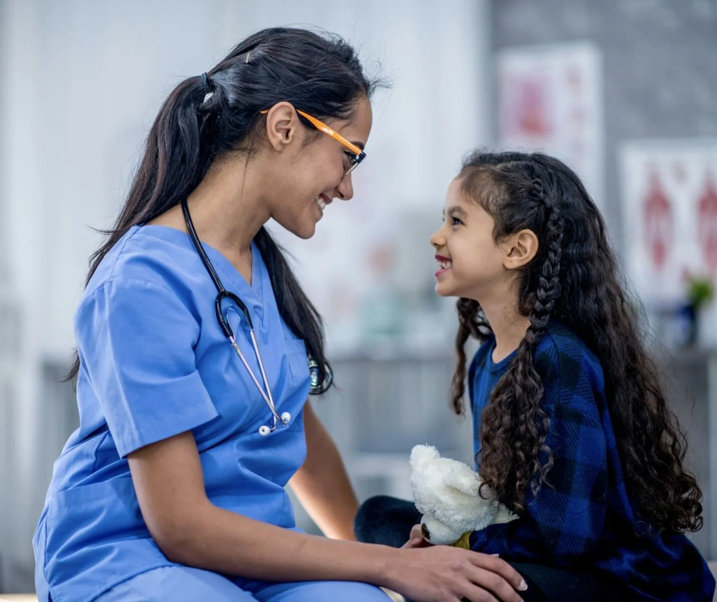 Smiling female pediatric therapist in blue scrubs interacting warmly with a young girl holding a white teddy bear.