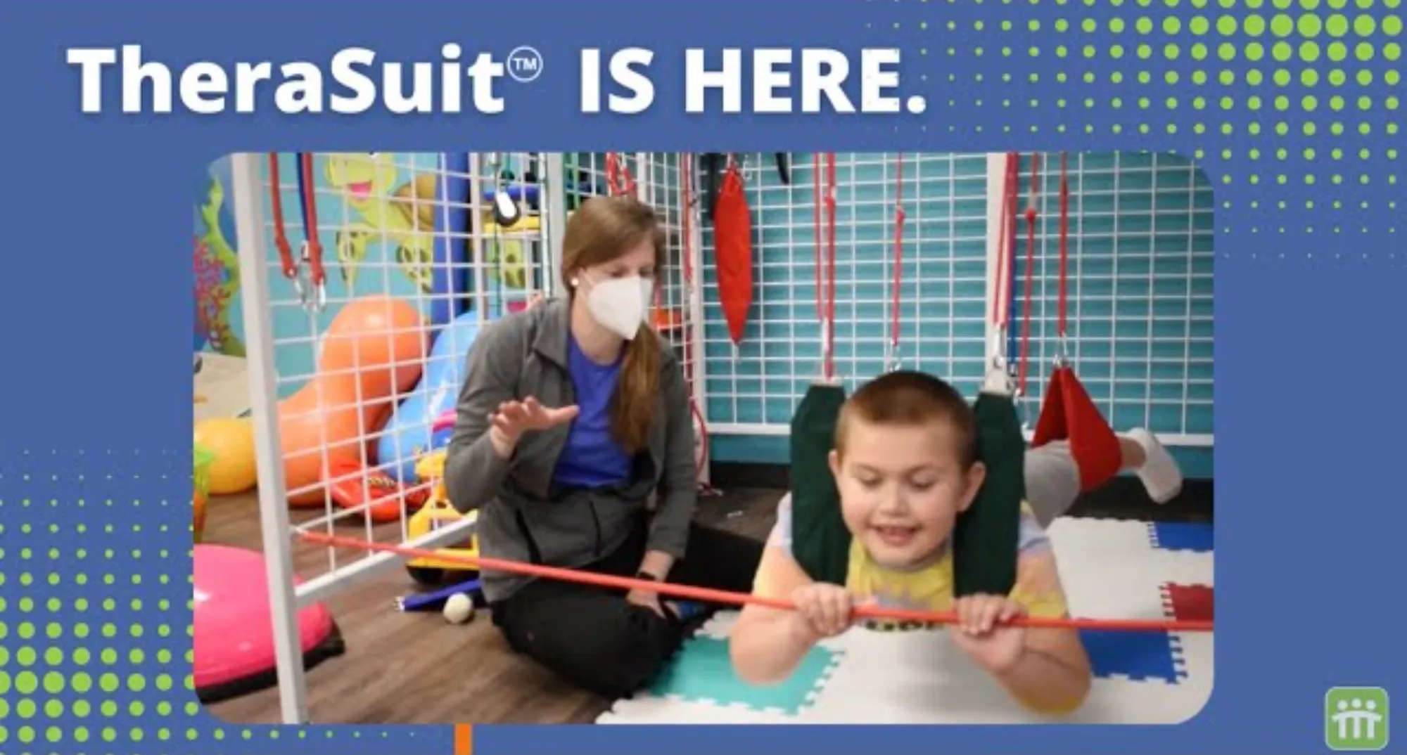Child suspended in a TheraSuit harness, holding a red rod, with a masked therapist guiding nearby in a therapy room.