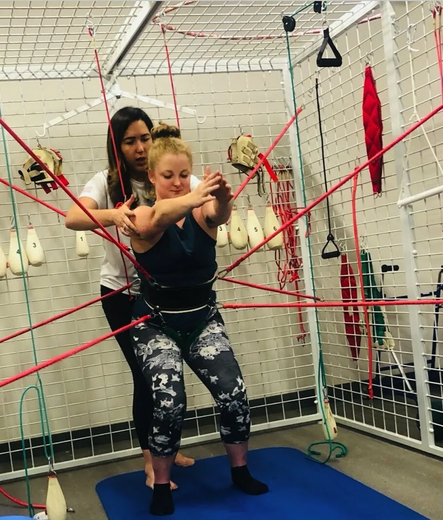 Therapist assists woman wearing a Therasuit with resistance bands performing a physical therapy exercise on a blue mat.