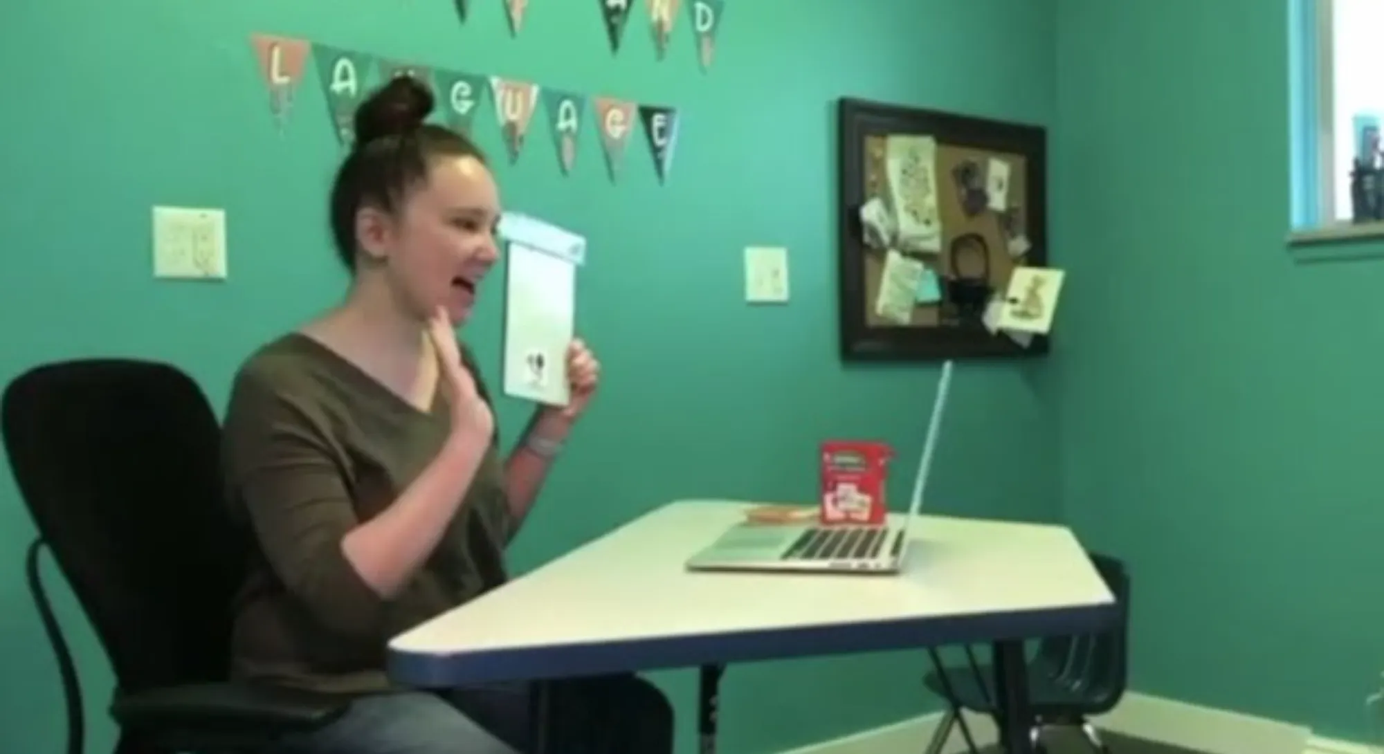 Woman sitting at a table in a teal room holding up a flashcard and gesturing during a virtual session on a laptop.
