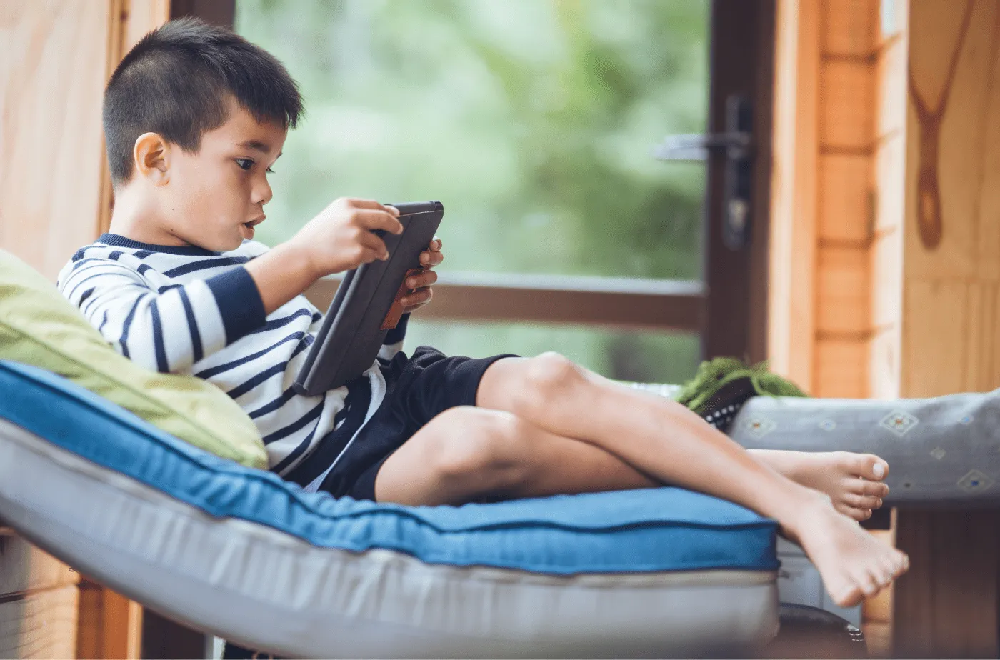 Young boy sitting on a cushioned chair indoors, focused on using a handheld tablet device.