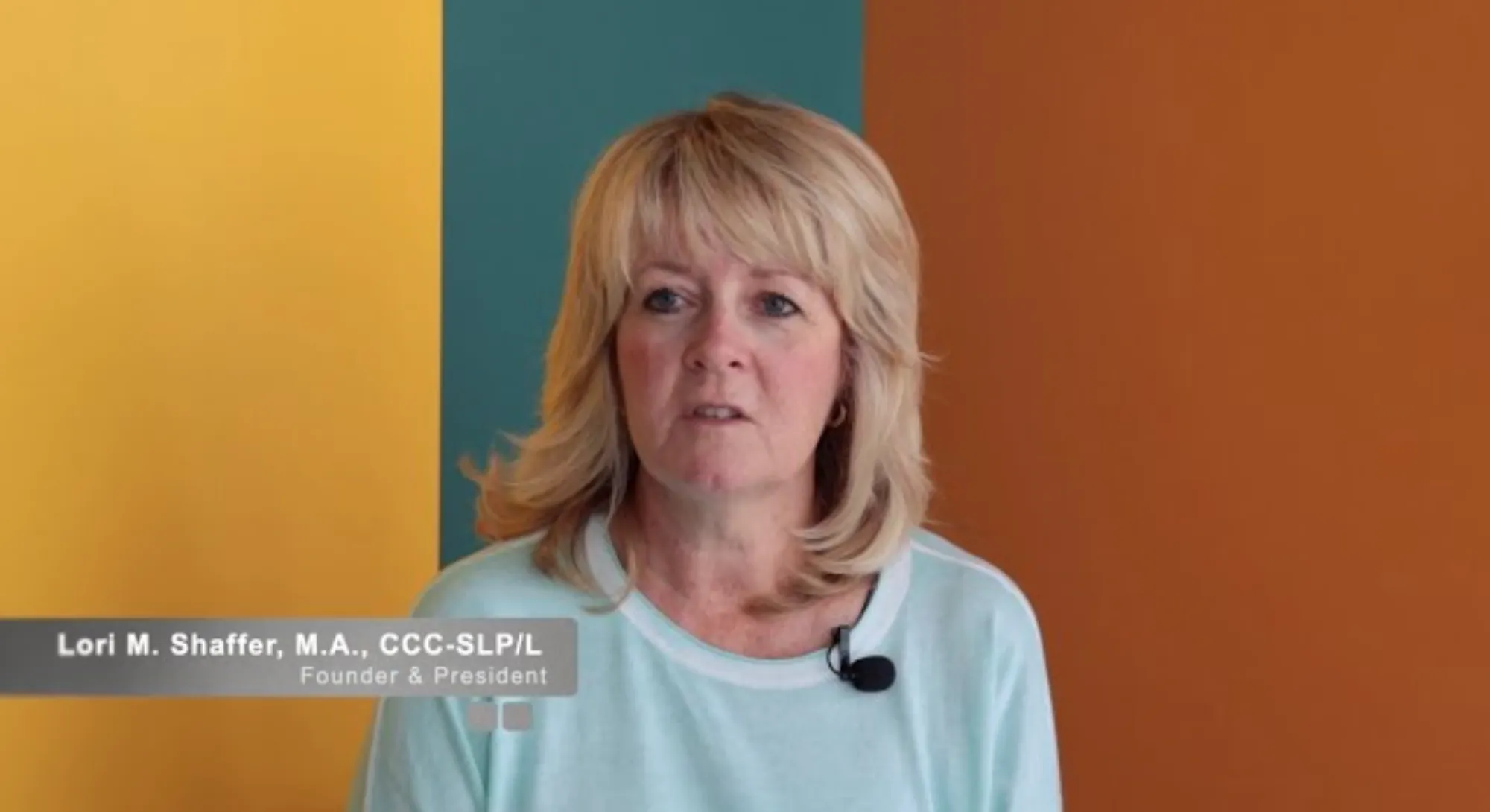 Medium shot of a woman with blonde hair wearing a light blue top and a microphone, standing against a background with three vertical color panels: yellow, teal, and orange.