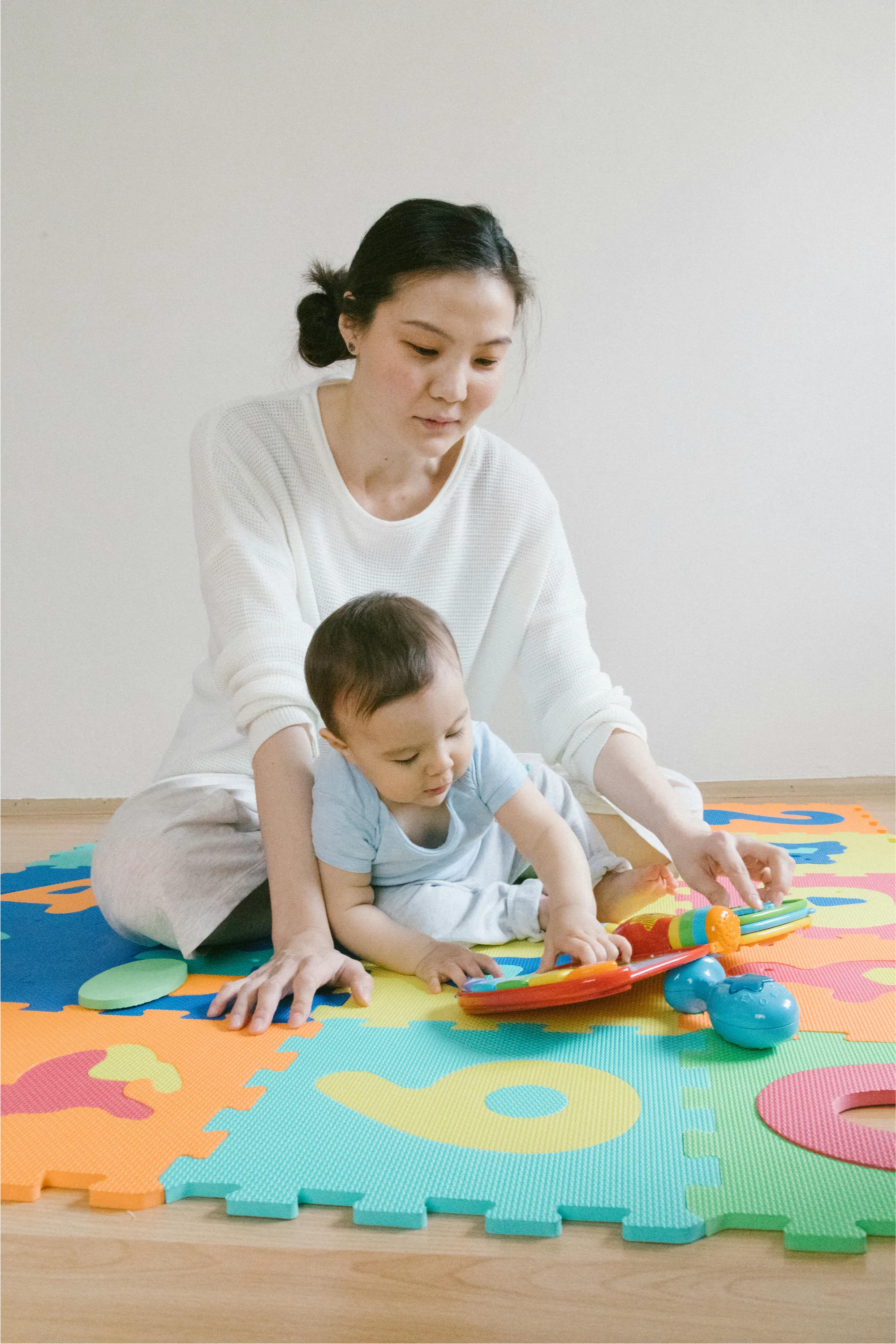Woman sitting on colorful foam alphabet mat playing with a baby who is touching a toy.