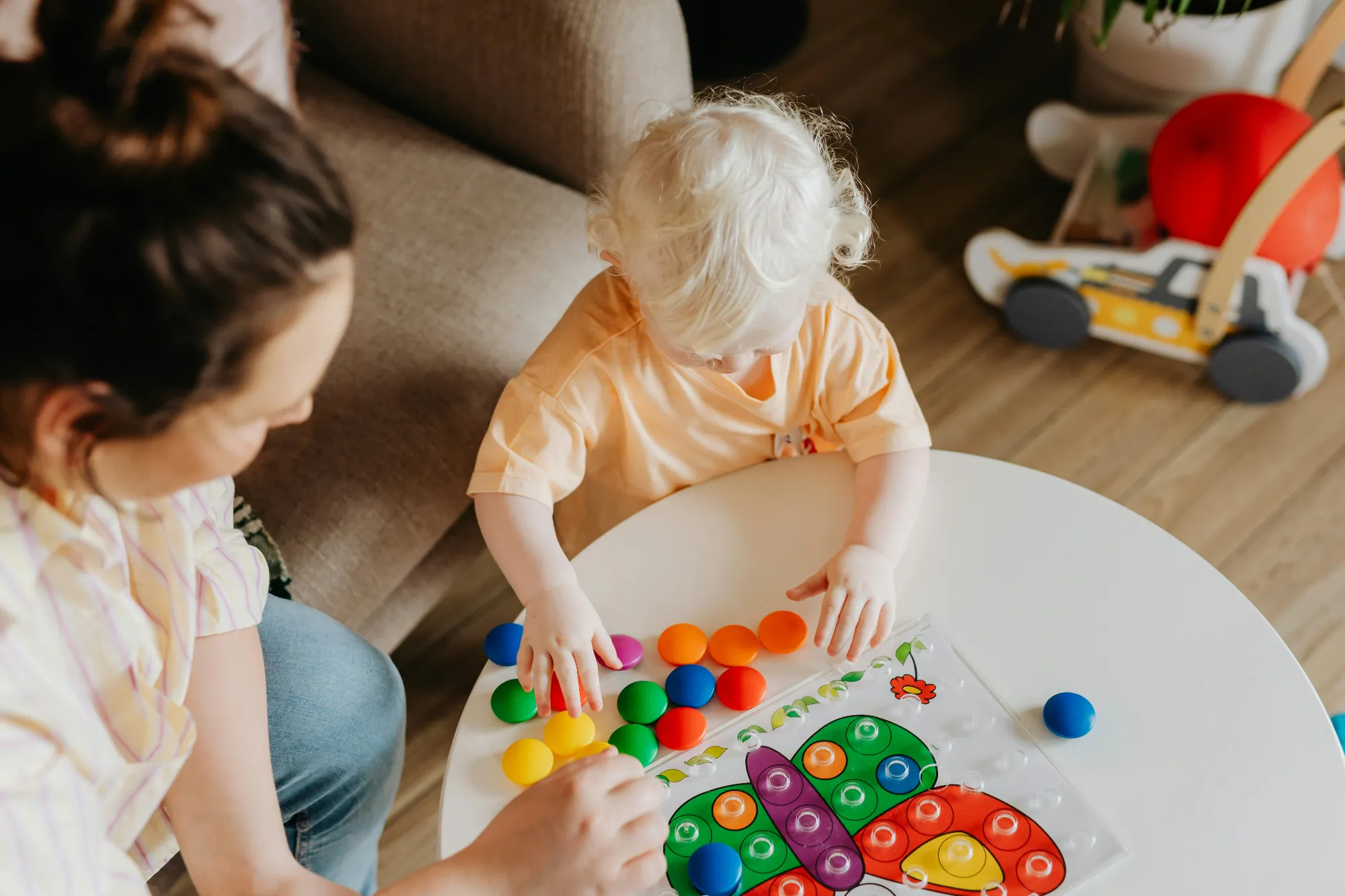 Young child with blonde hair playing with colorful pegboard butterfly toy at a white table with an adult nearby.