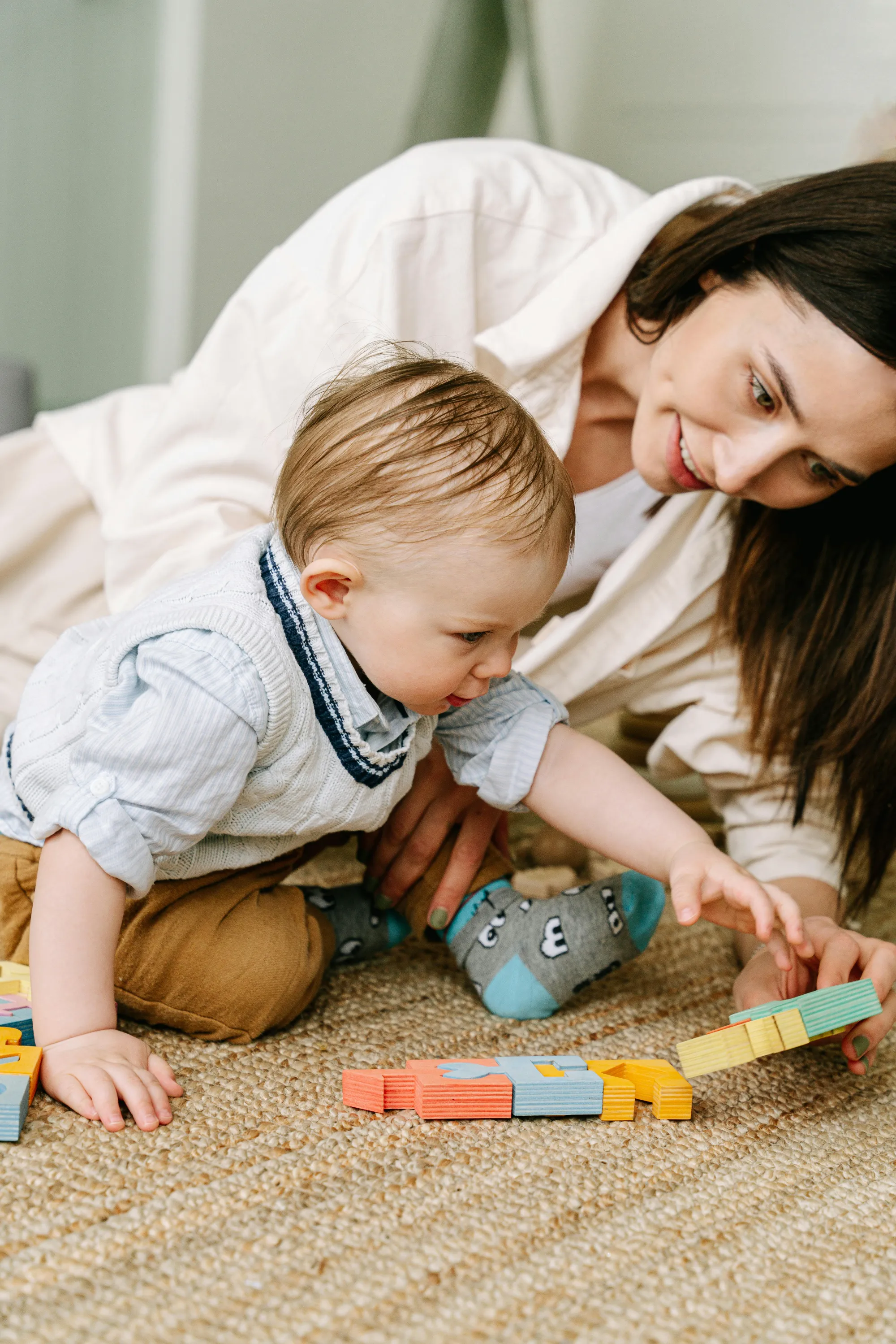 A woman and a toddler play together on a textured rug, stacking colorful wooden blocks.