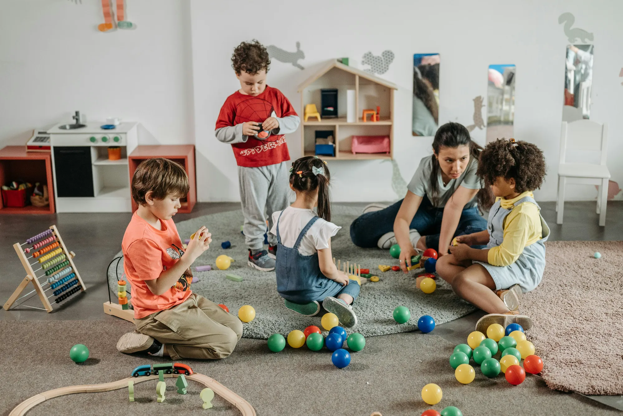 A woman interacts and plays with four children surrounded by colorful balls and toys on a carpet in a playroom.