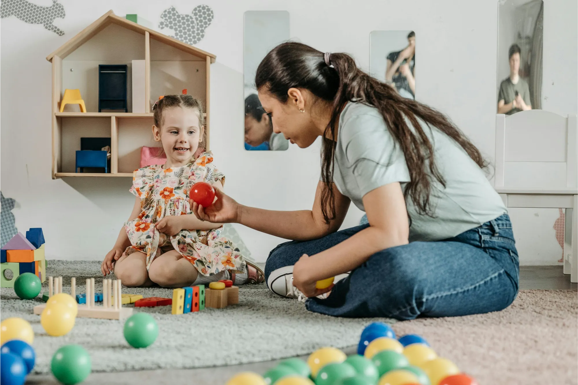 Woman playing with a smiling young girl on a carpet surrounded by colorful toy balls and wooden blocks in a playroom.