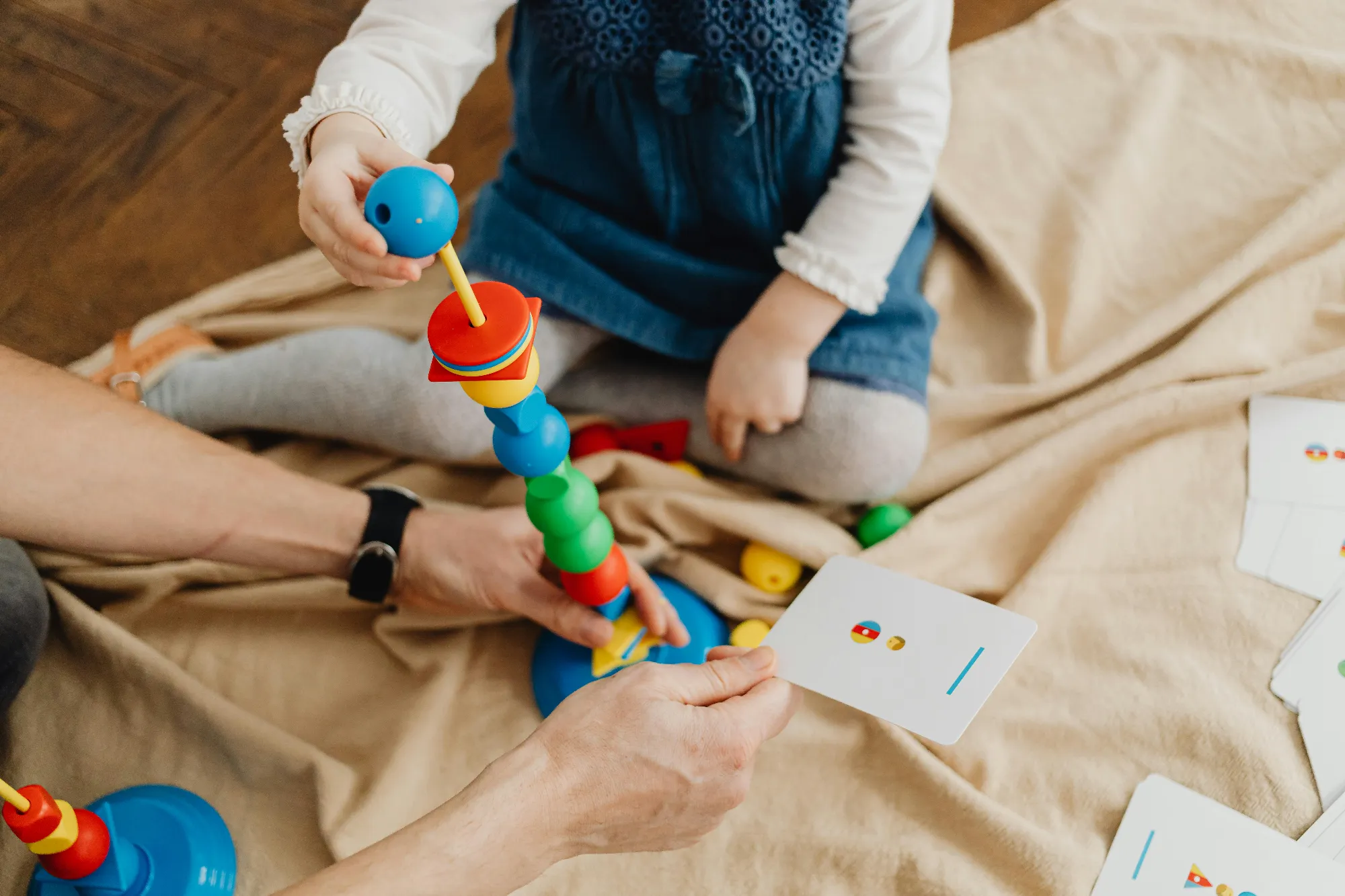 Child playing with a colorful stacking toy while an adult holds a matching activity card nearby.