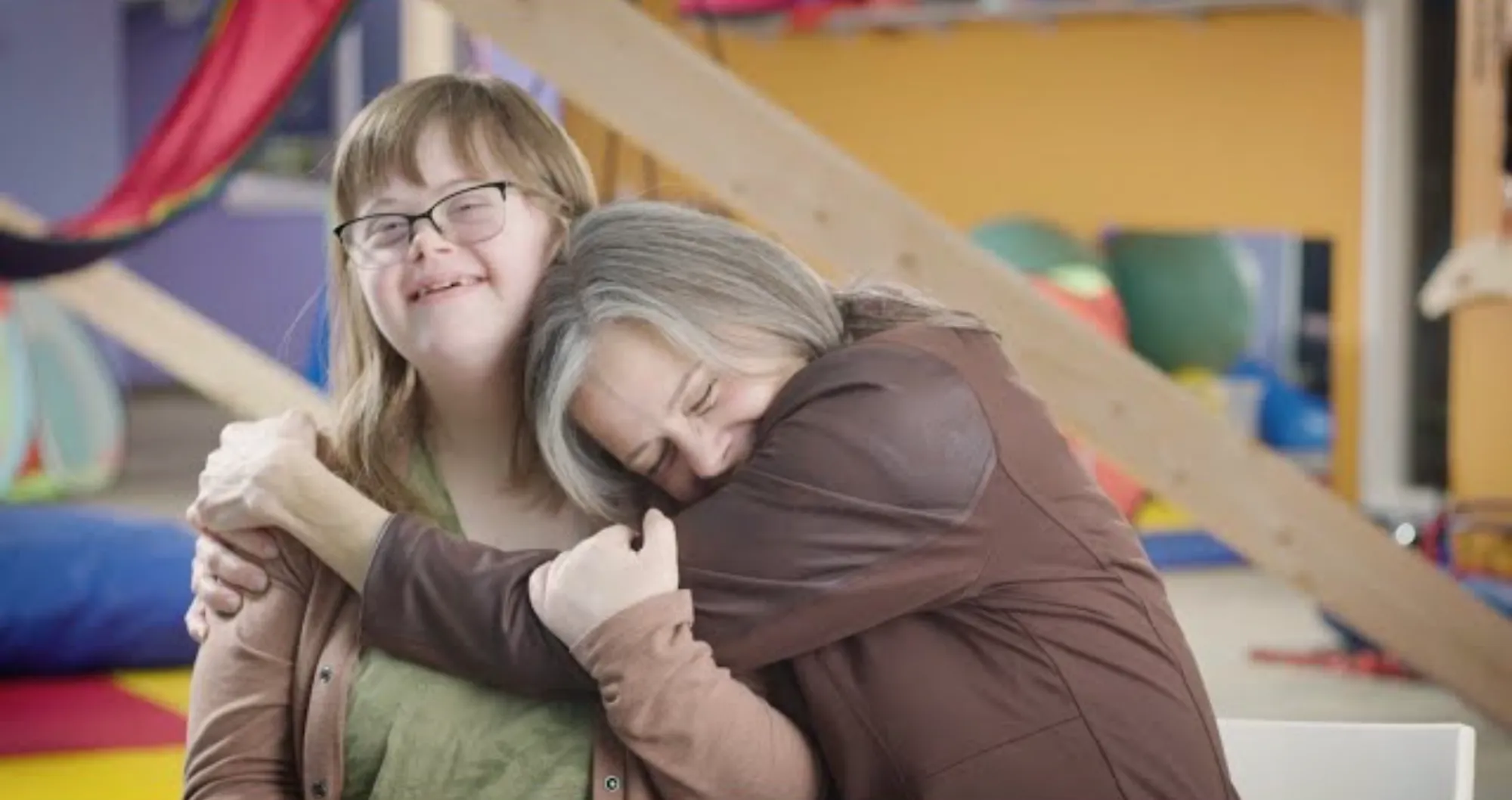Smiling young woman with glasses and Down syndrome embracing an older woman who hugs her tightly with eyes closed.