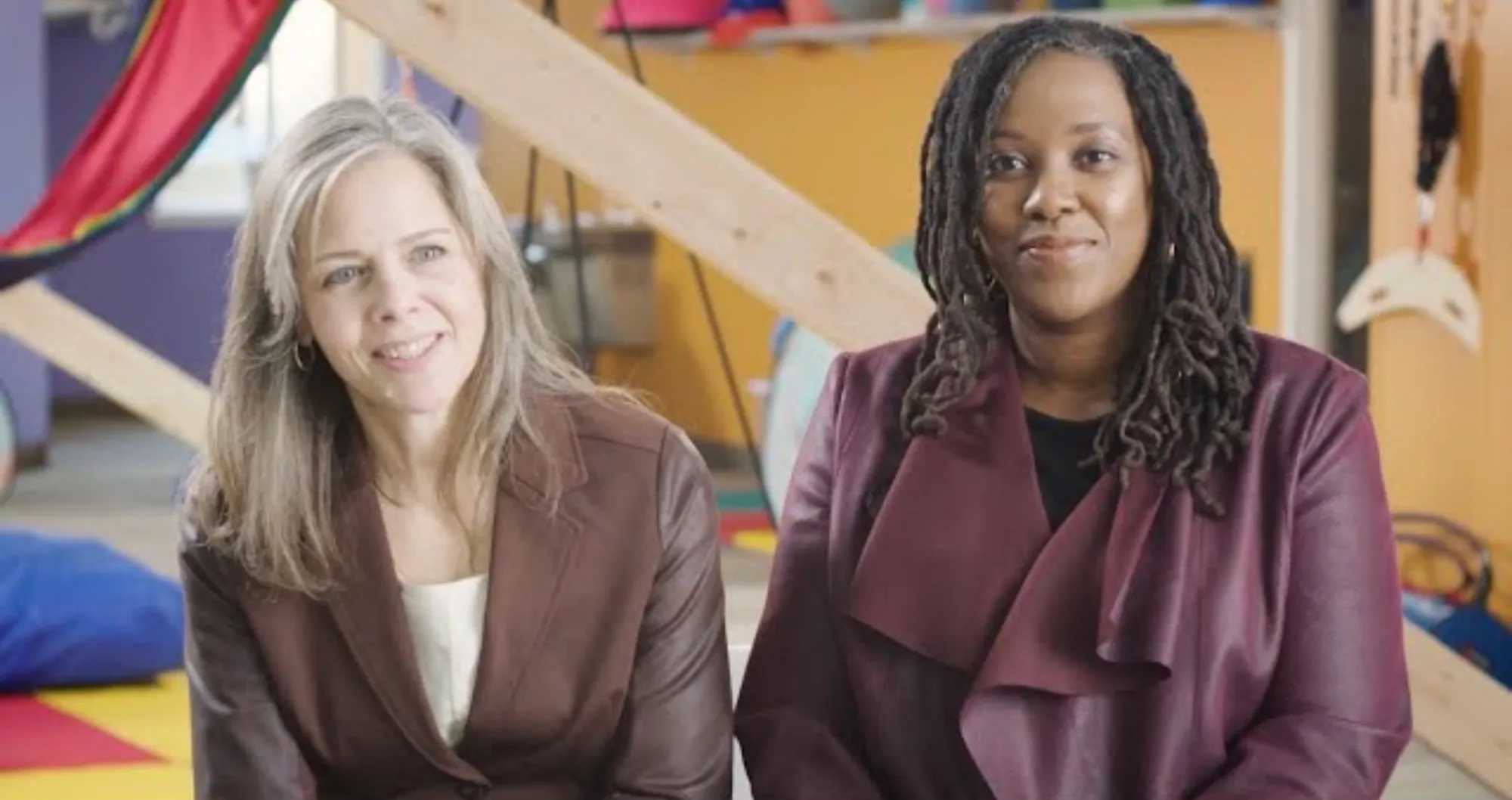 Two women sitting indoors in a colorful room with wooden beams in the background, both wearing brown jackets and smiling.