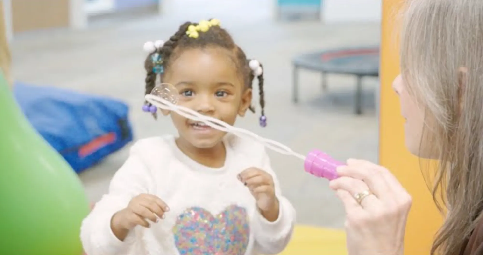 Young girl with colorful hair beads playing with bubbles being blown by an adult woman.