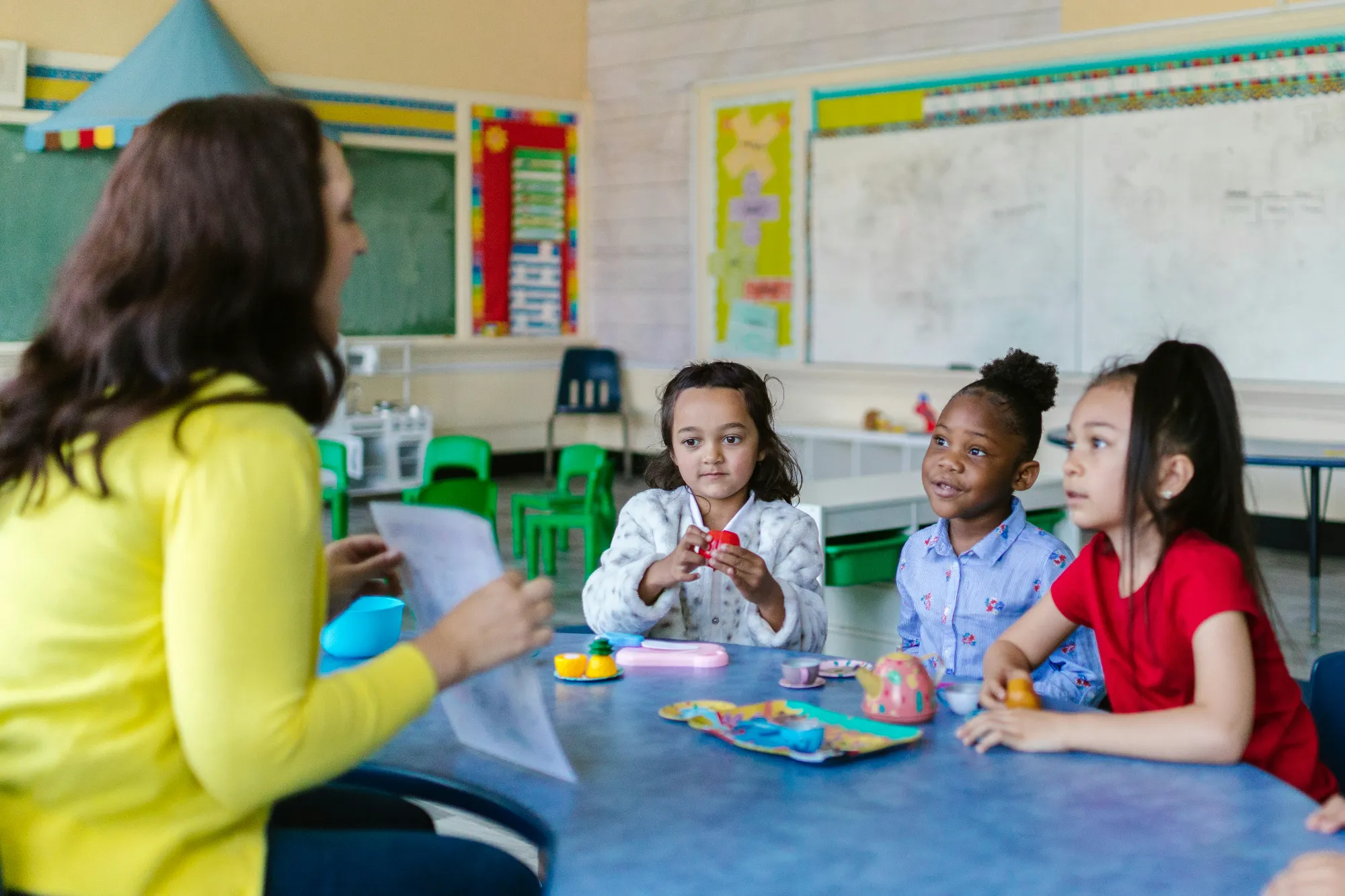 Teacher in yellow sweater engaging with three young children sitting at a table with toy tea set in a classroom.