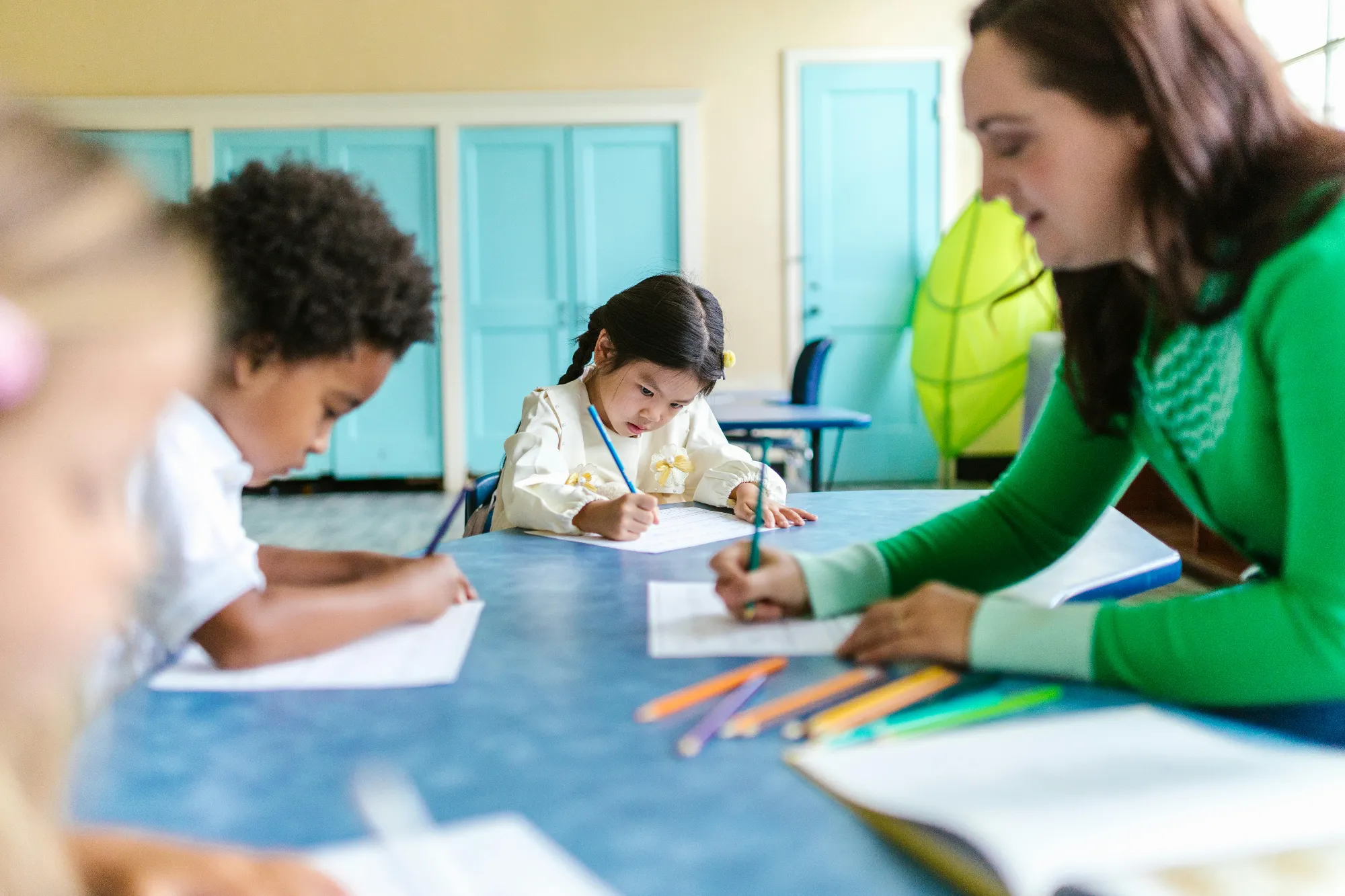 Teacher and young children focused on writing at a classroom table with colorful pencils nearby.