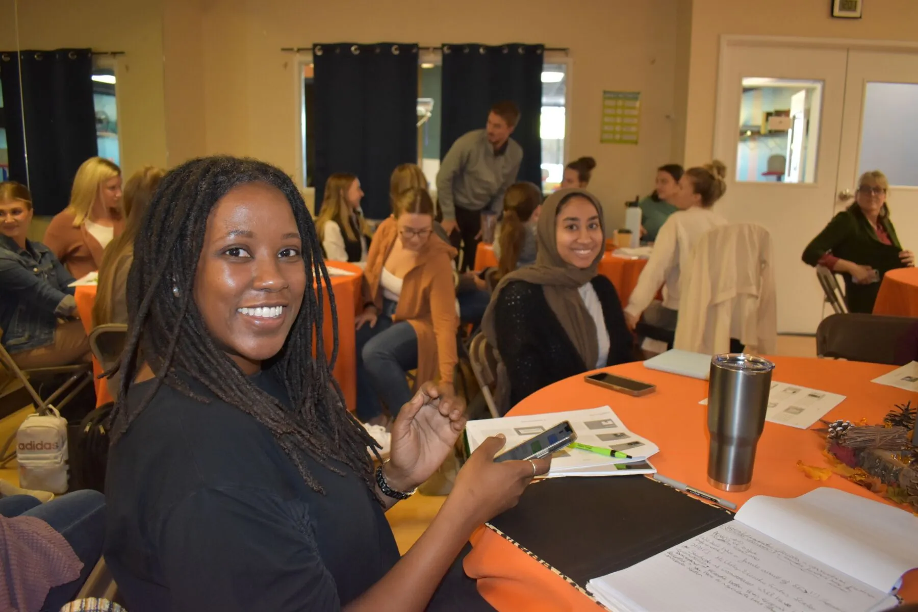 Smiling woman with dreadlocks holding a smartphone at a table with notebooks and a stainless steel tumbler in a group meeting.