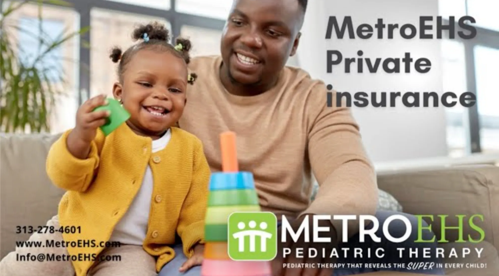 A smiling toddler in a yellow cardigan plays with colorful stacking rings while an adult man watches and smiles nearby.