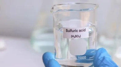 A gloved hand holding a glass beaker filled with liquid labeled Sulfuric acid H2SO4.