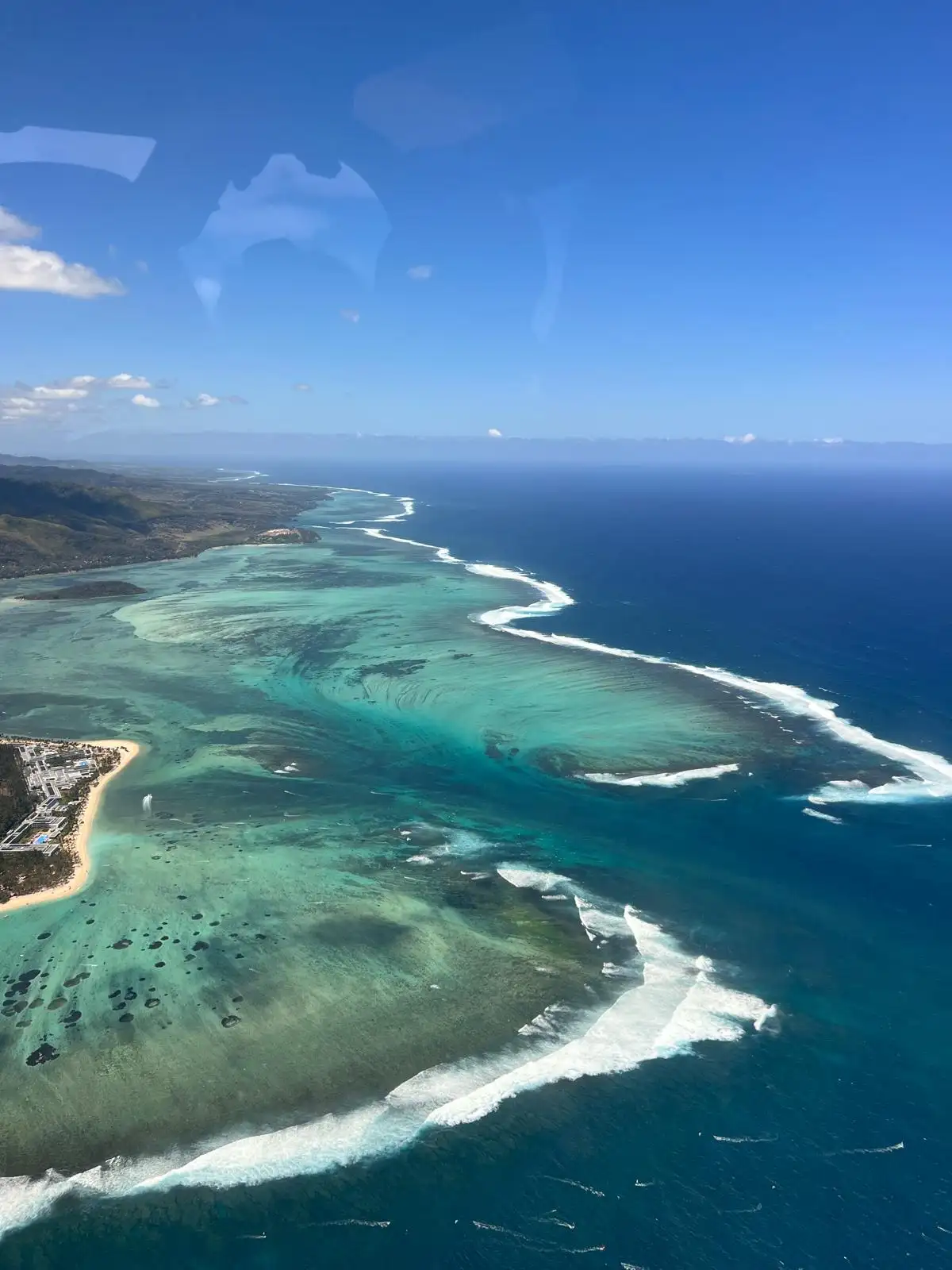 Vue aérienne d'une côte tropicale avec une eau turquoise, une plage de sable et des vagues blanches déferlantes sous un ciel bleu clair.