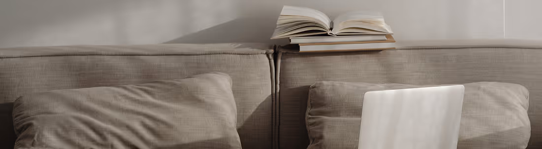 Hellgraues Sofa mit zwei passenden Kissen, einem aufgeklappten Buch und einem Laptop daneben in einem hellen Raum.