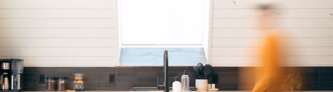 Moderne Küchenspüle mit schwarzem Wasserhahn unter einem großen Fenster, umgeben von einer Kaffeemaschine, Gläsern und Utensilien, mit einer verschwommenen Person in einem orangefarbenen Hemd auf der rechten Seite.