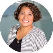 Lächelnde Frau mit lockigem kurzen Haar, gestreiftem Blazer und schwarzem Oberteil vor verschwommenem Hintergrund.