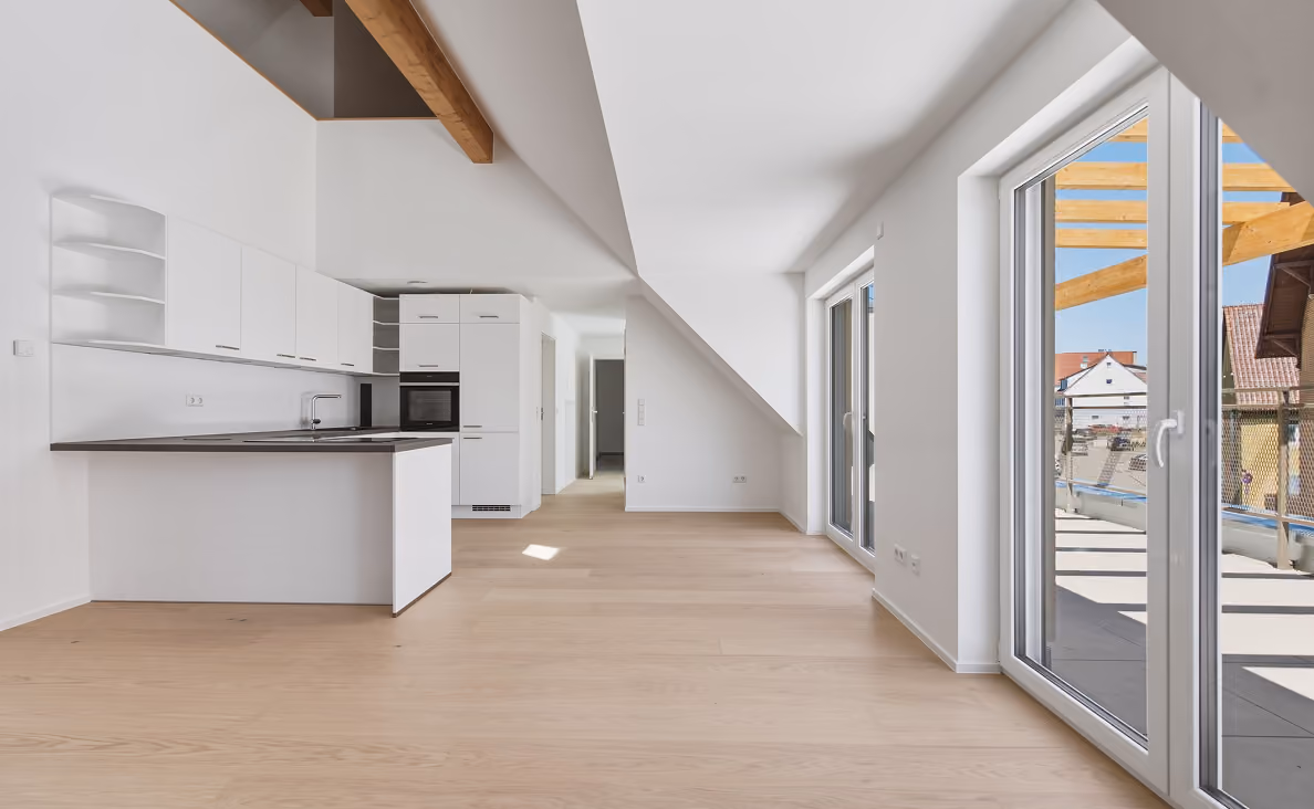 Helle, moderne Wohnung mit offener Küche, weißen Schränken, Holzbalken an der Decke und großen Fenstern, die auf einen Balkon mit Blick auf Nachbarhäuser führen.