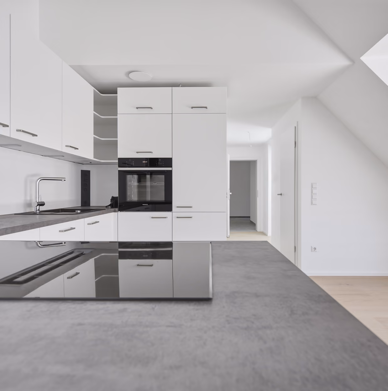 Moderne, minimalistische weiße Küche mit grauer Arbeitsplatte, eingebautem schwarzen Backofen, Induktionskochfeld und Spüle, offenes Regal und heller Flur im Hintergrund.