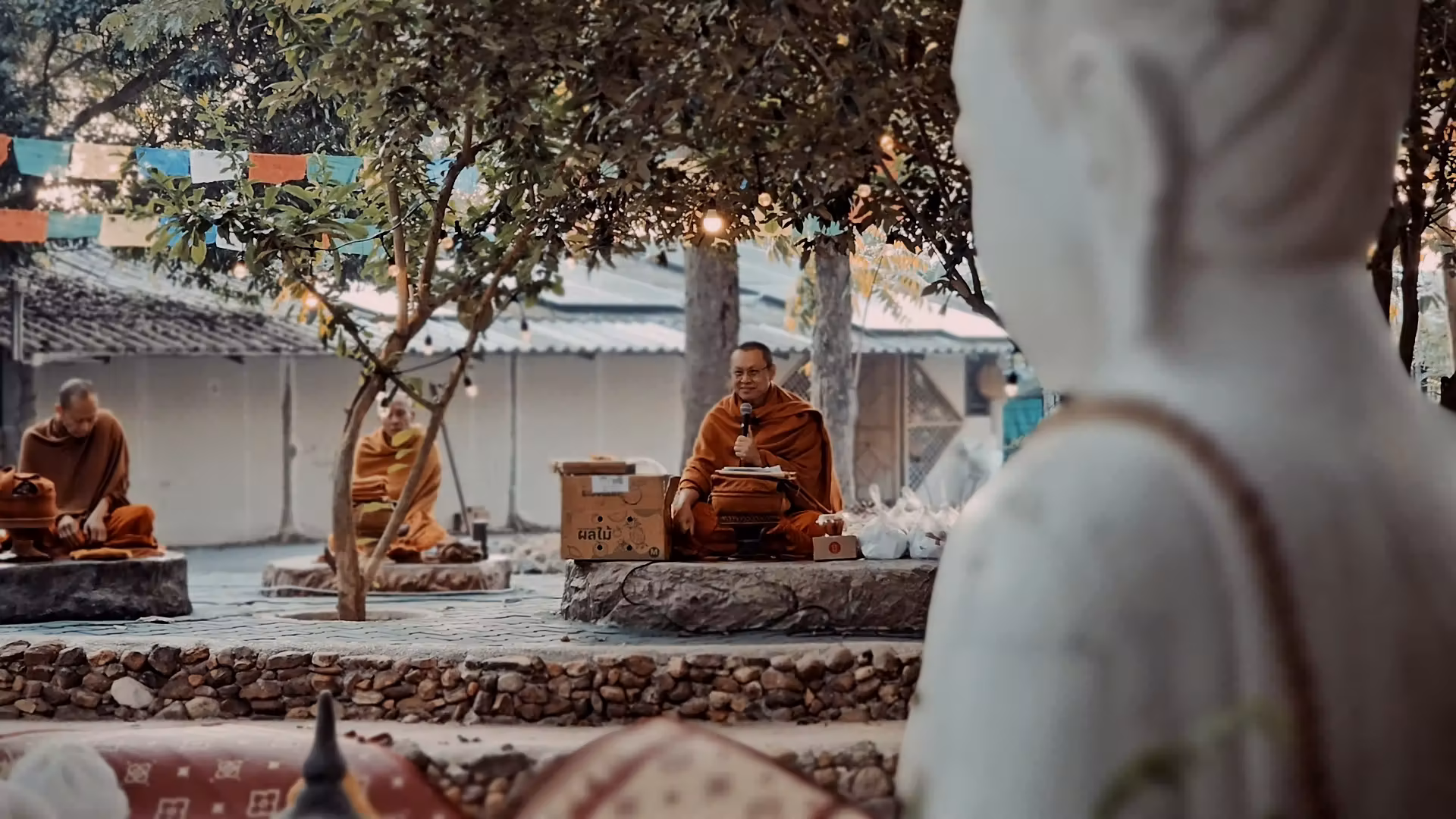 Three Buddhist monks in orange robes sitting outdoors on stone platforms under trees, with one monk holding a microphone.