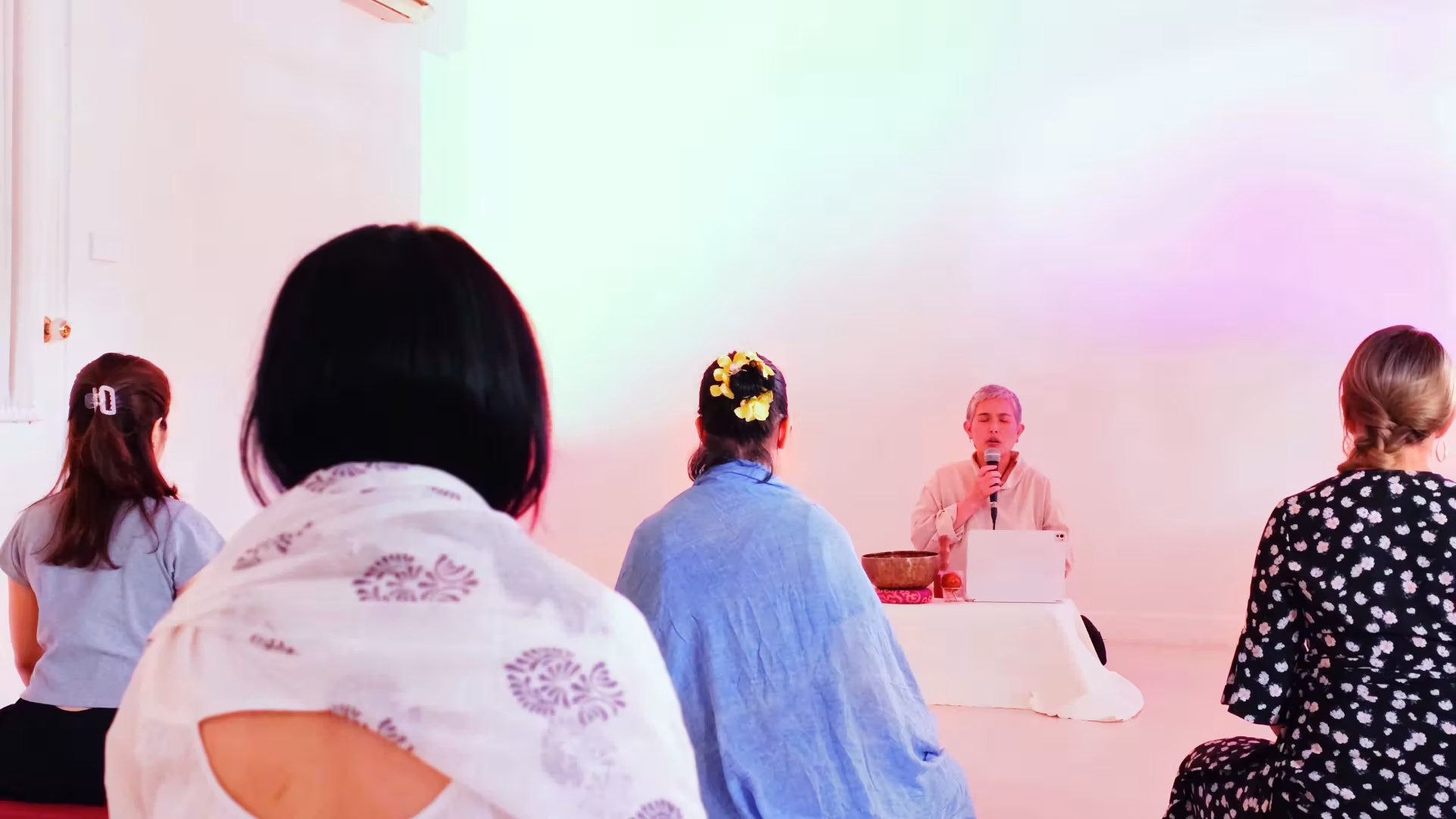 Group of five people sitting cross-legged indoors, facing a person with a microphone leading a meditation or mindfulness session.